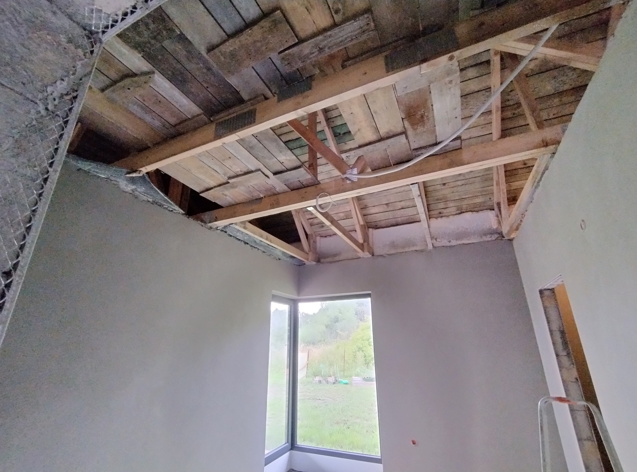 Surowe wnętrze z oknem: świeżo otynkowane ściany, drewniany strop z widoczną konstrukcją, fragment siatki na ścianie. W tle zieleń za oknem.