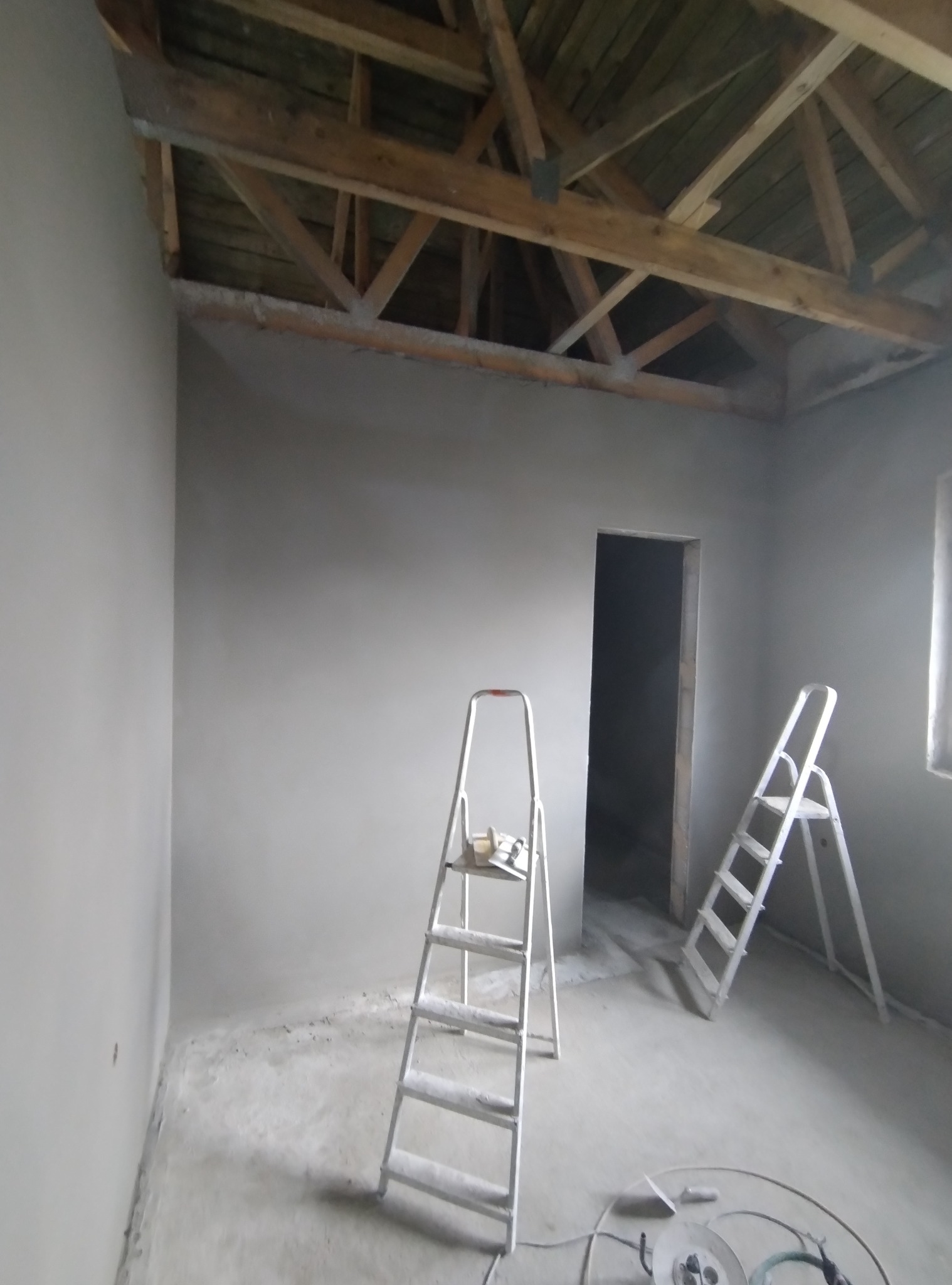 Wnętrze po tynkowaniu: gładkie, szare ściany, drewniana więźba dachowa, dwie drabiny malarskie i narzędzia na podłodze. Widok na otwór drzwiowy.