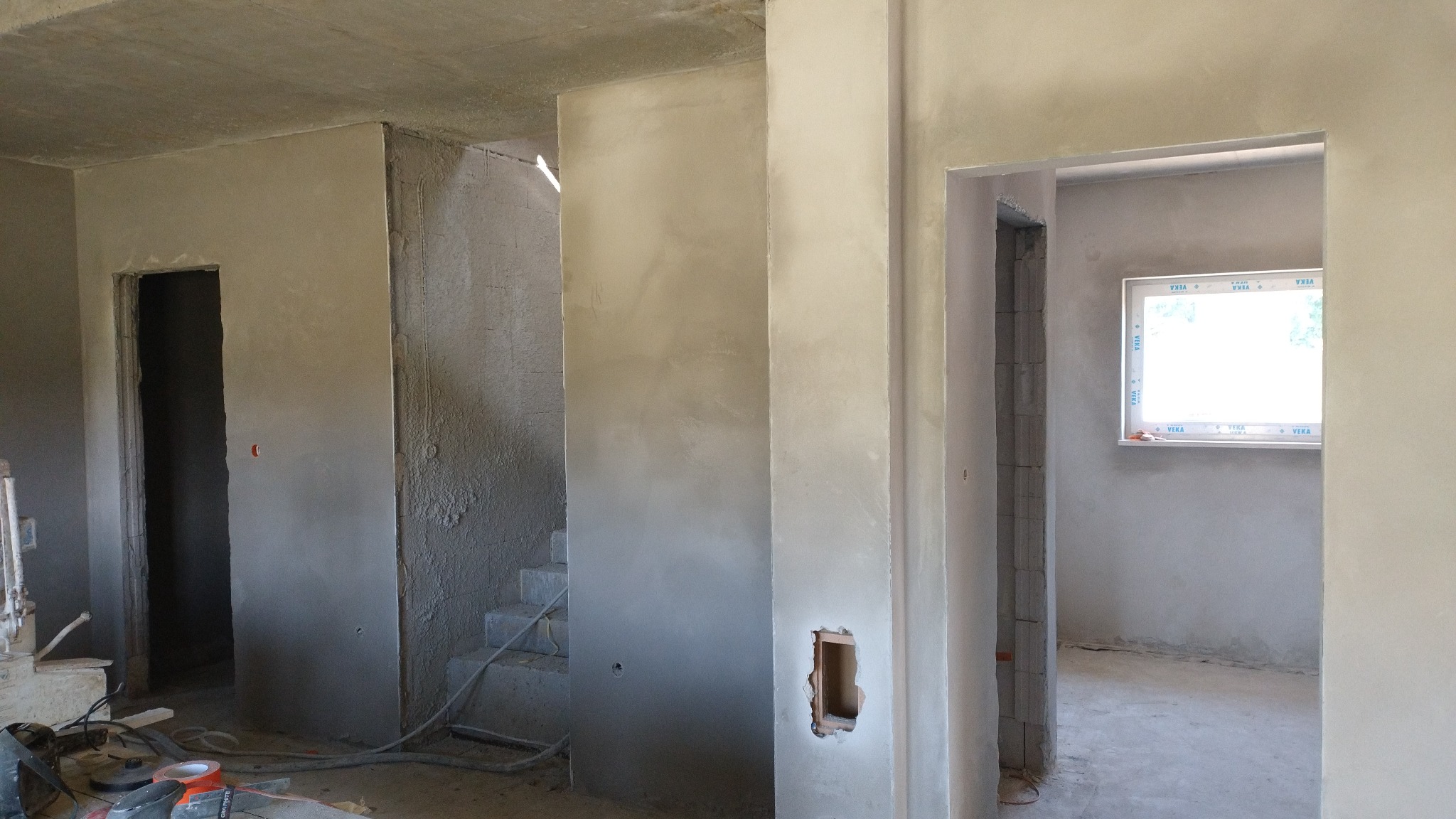 Wnętrze w trakcie tynkowania: surowe ściany z widocznymi śladami tynku, otwory drzwiowe i okienne, narzędzia na podłodze. Widoczne schody w głębi pomieszczenia.