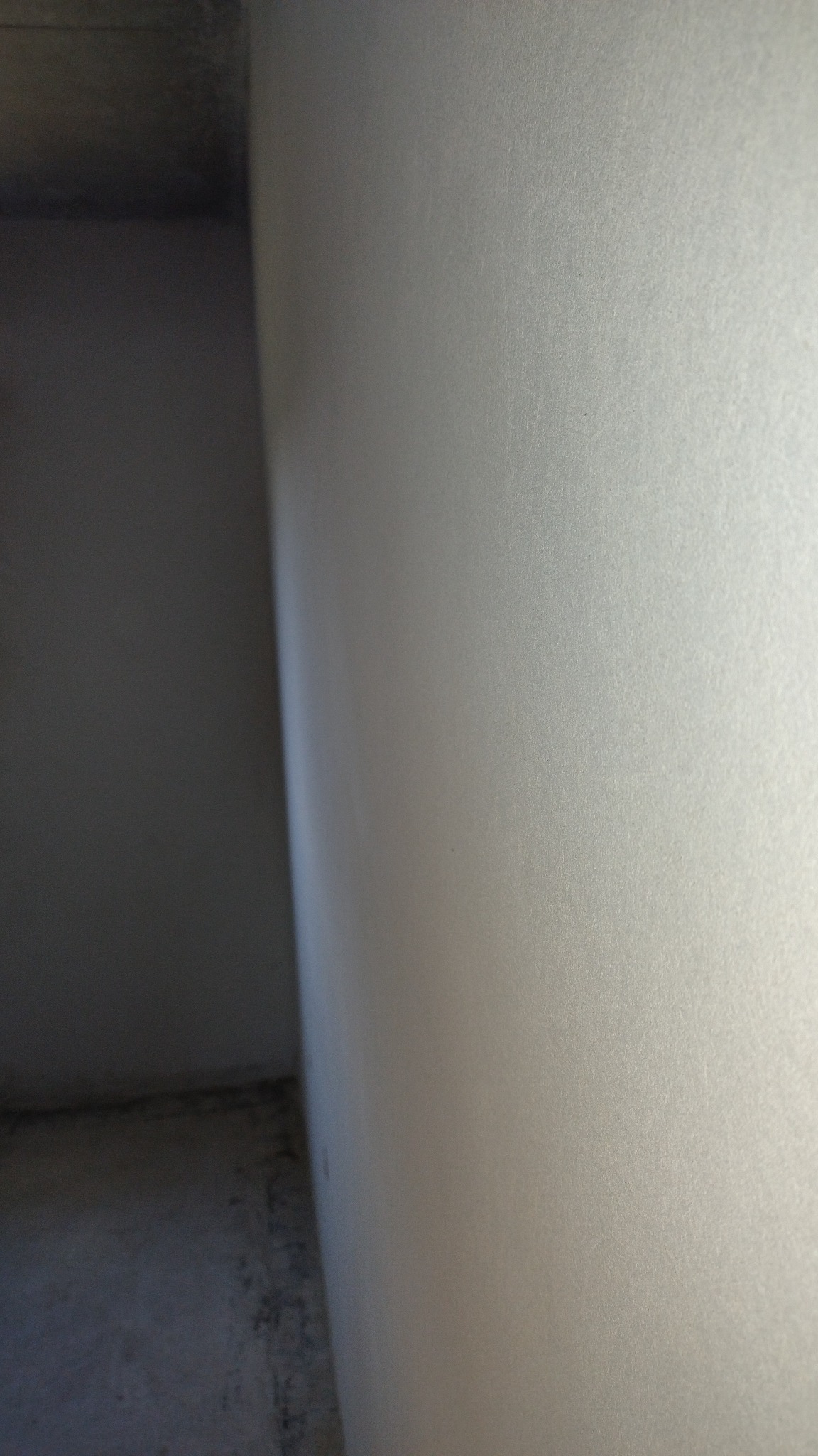 Gładka, jasna ściana po tynkowaniu maszynowym w rogu pomieszczenia. Widoczny fragment sufitu i podłogi w surowym stanie. Minimalistyczne ujęcie.