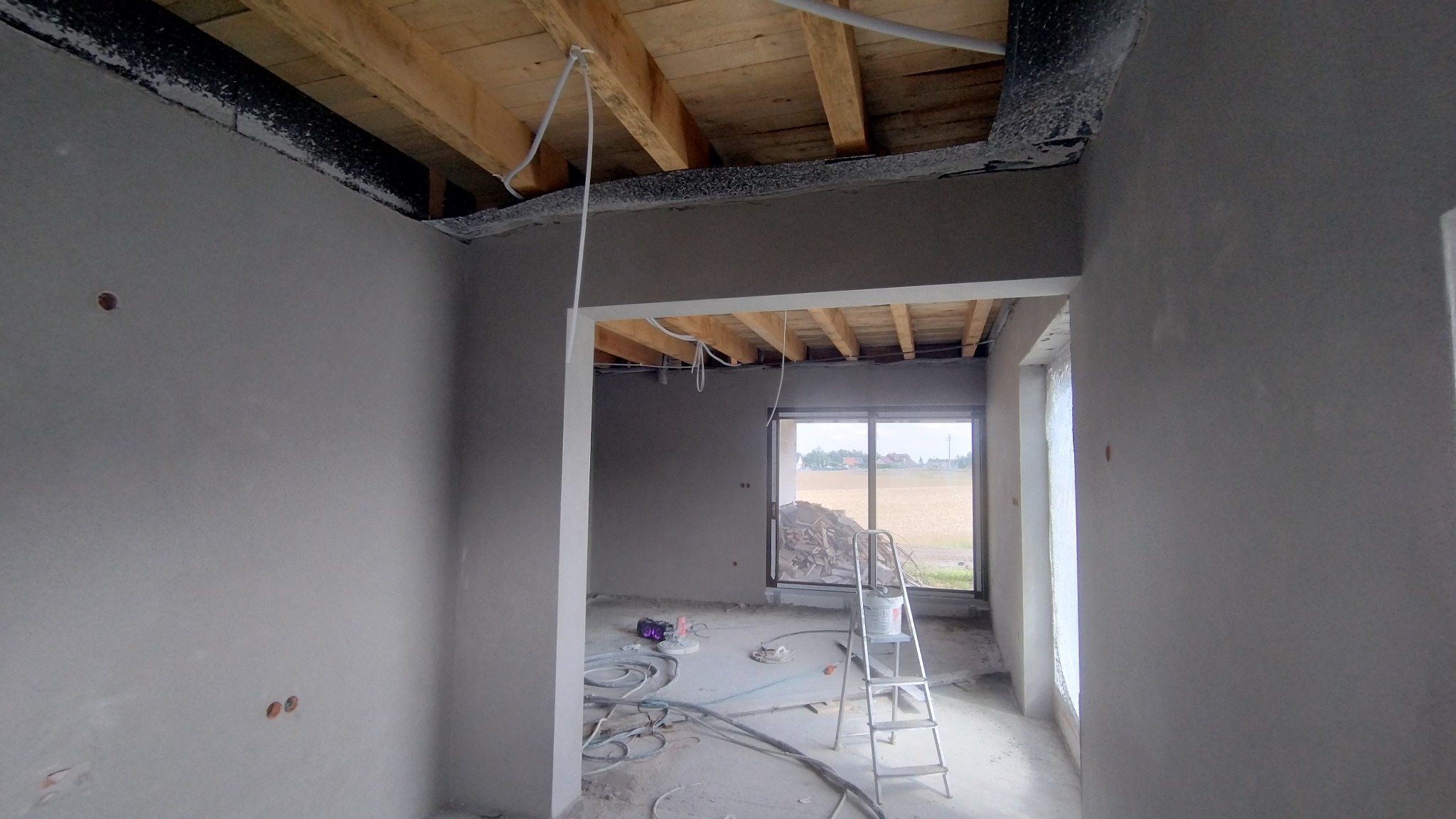 Wnętrze w stanie surowym po tynkowaniu ścian. Widoczne drewniane elementy stropu, instalacje elektryczne, okno z widokiem na pole i drabina malarska.