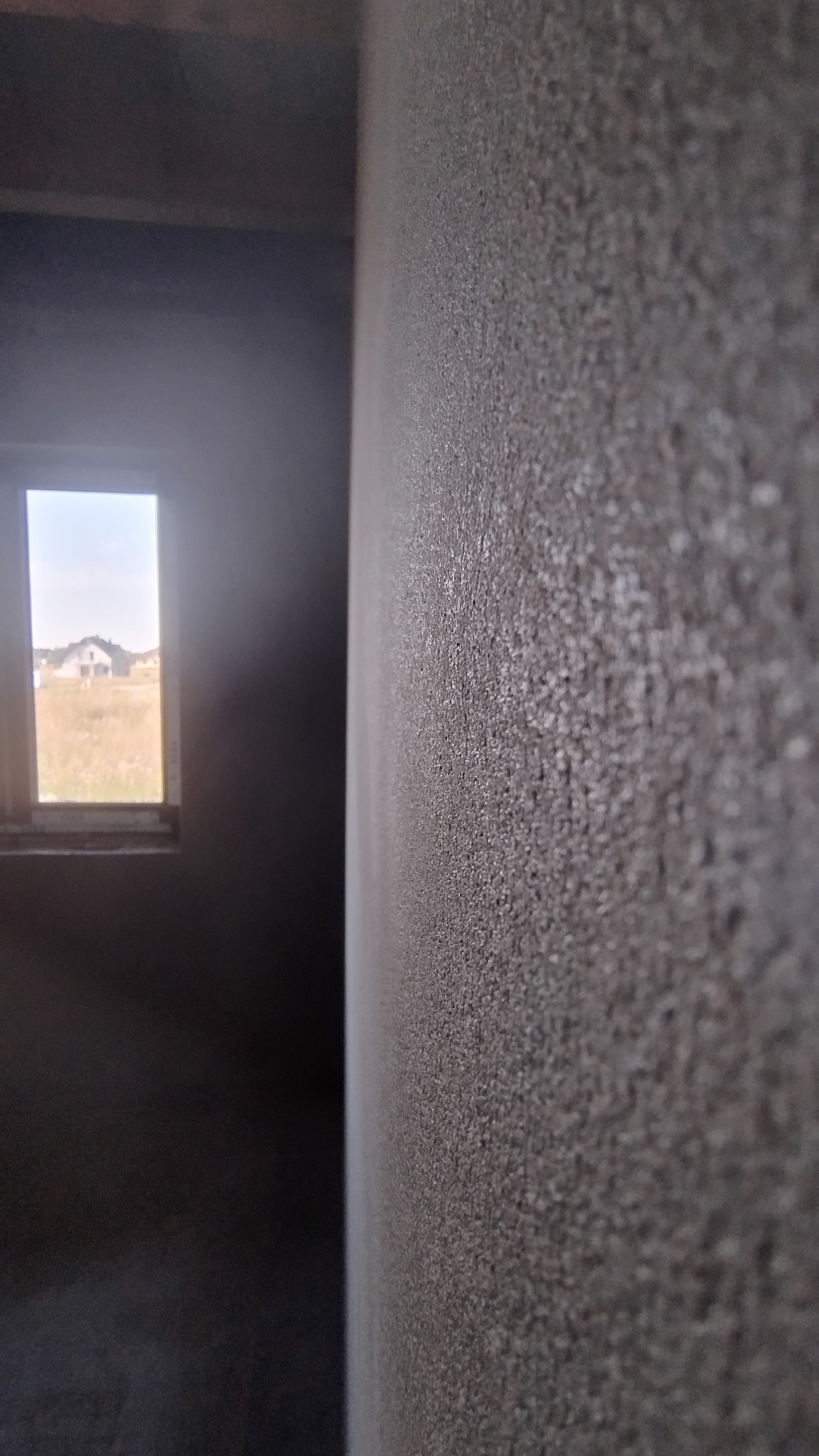 Ściana wewnętrzna z nałożonym tynkiem strukturalnym, widok z bliska. W tle okno z widokiem na budynek i pole. Surowy stan wykończenia wnętrza.
