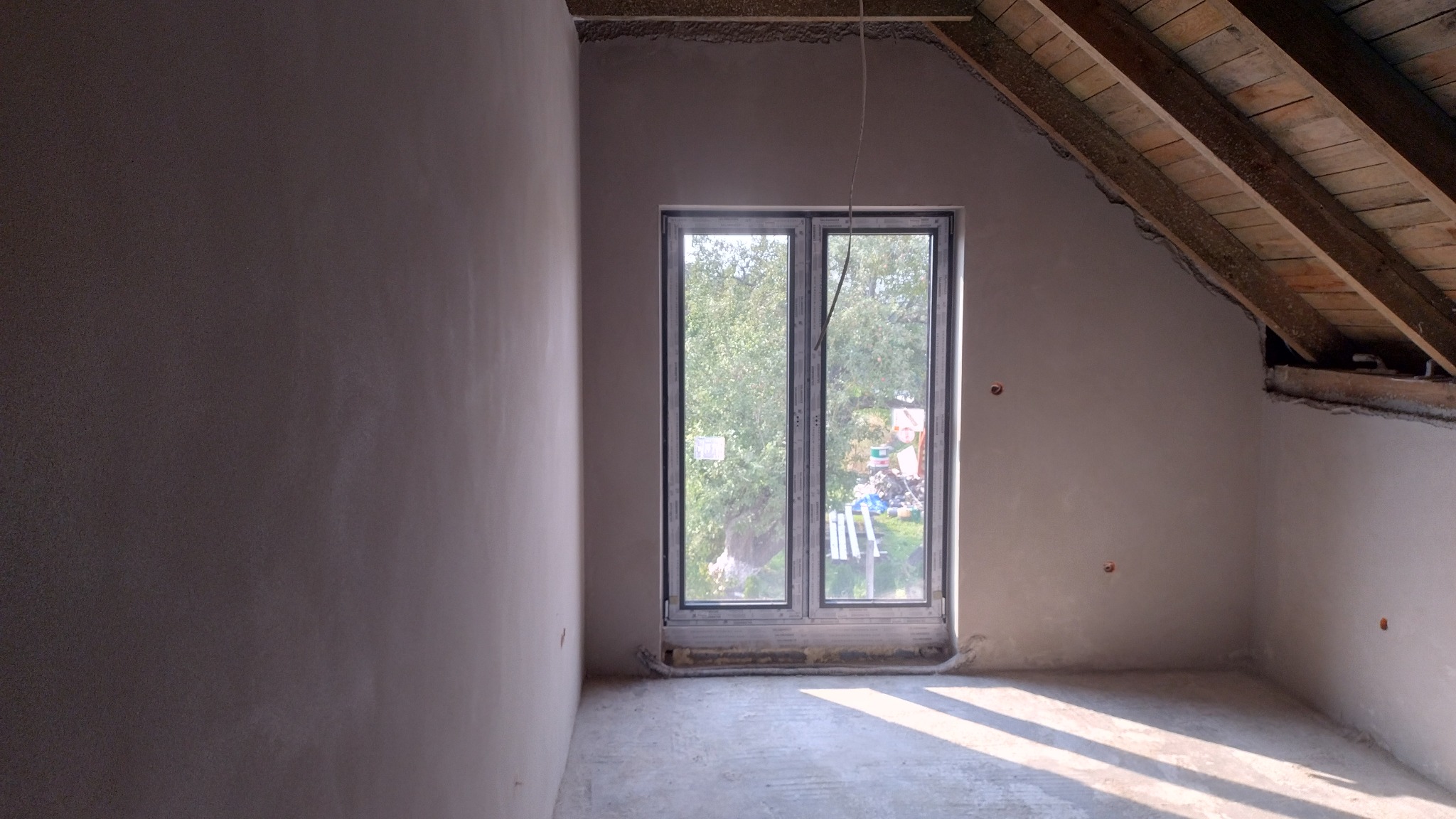 Pokój na poddaszu po tynkowaniu ścian, widoczne surowe drewno więźby dachowej i okno z widokiem na zieleń. Ściany gładkie, przygotowane do dalszych prac wykończeniowych.
