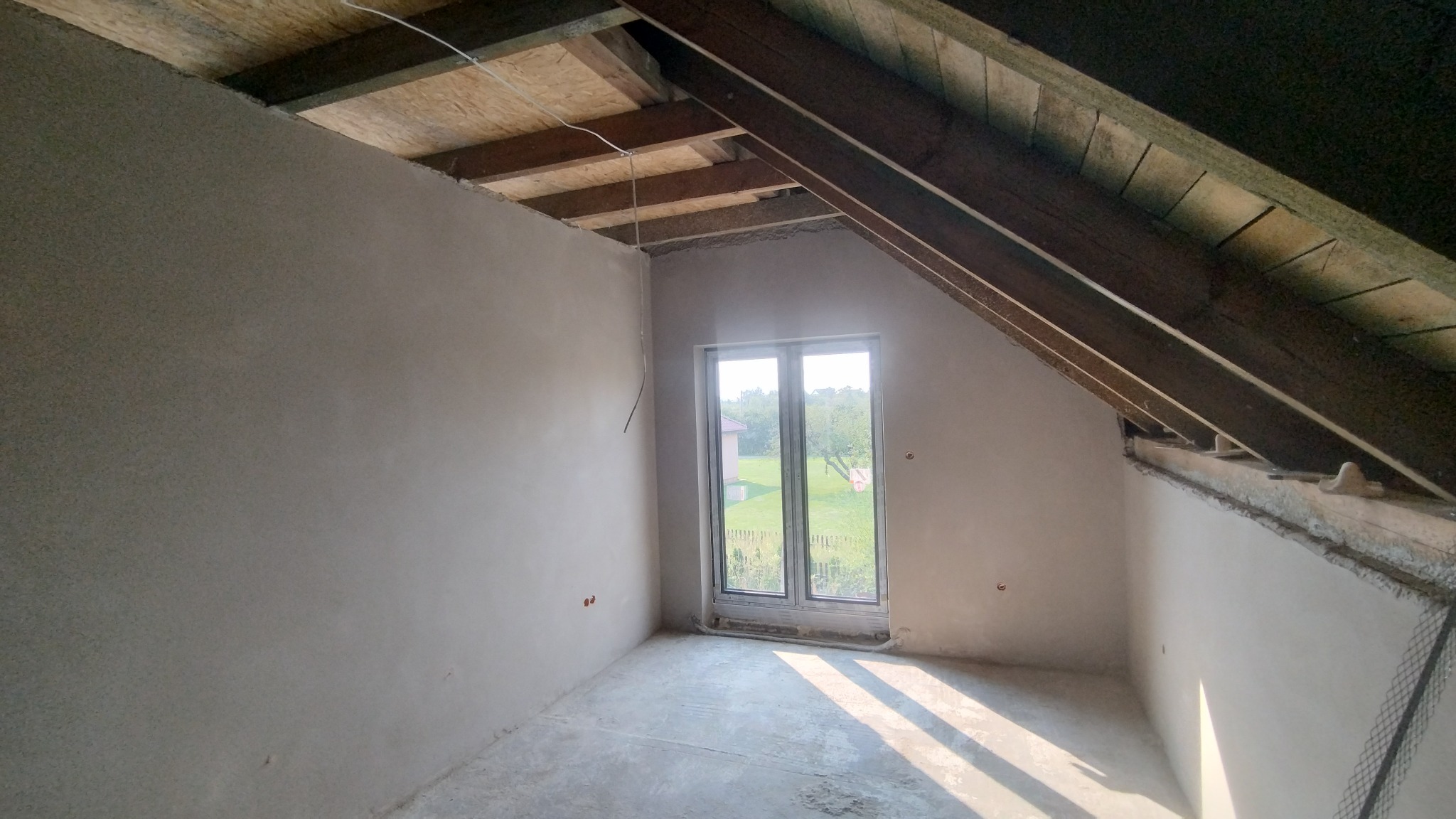 Pokój na poddaszu po tynkowaniu, widoczne surowe drewno konstrukcji dachu, okno z widokiem na zieleń. Ściany gładkie, przygotowane do dalszych prac wykończeniowych.
