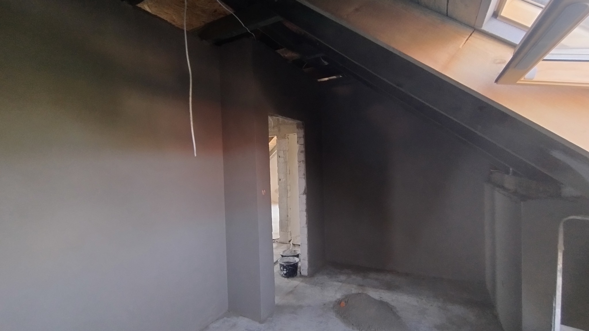 Szare, świeżo otynkowane poddasze z oknem dachowym i widocznym fragmentem konstrukcji. W tle widoczne wiadro i przejście do innego pomieszczenia.