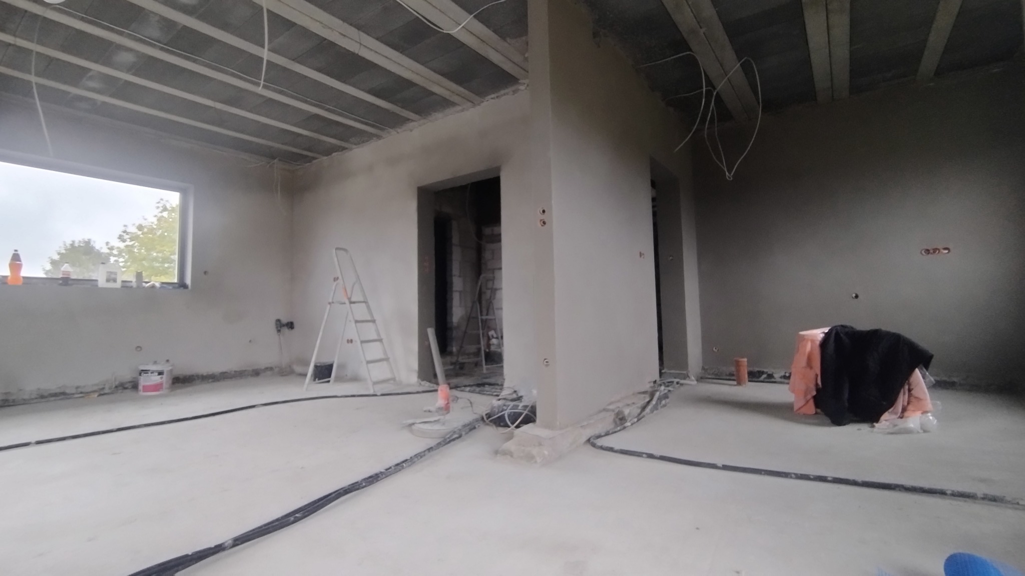 Wnętrze w trakcie tynkowania: świeżo otynkowane ściany, drabina, kable na podłodze, okno z widokiem na zewnątrz. Widoczne ślady prac tynkarskich.