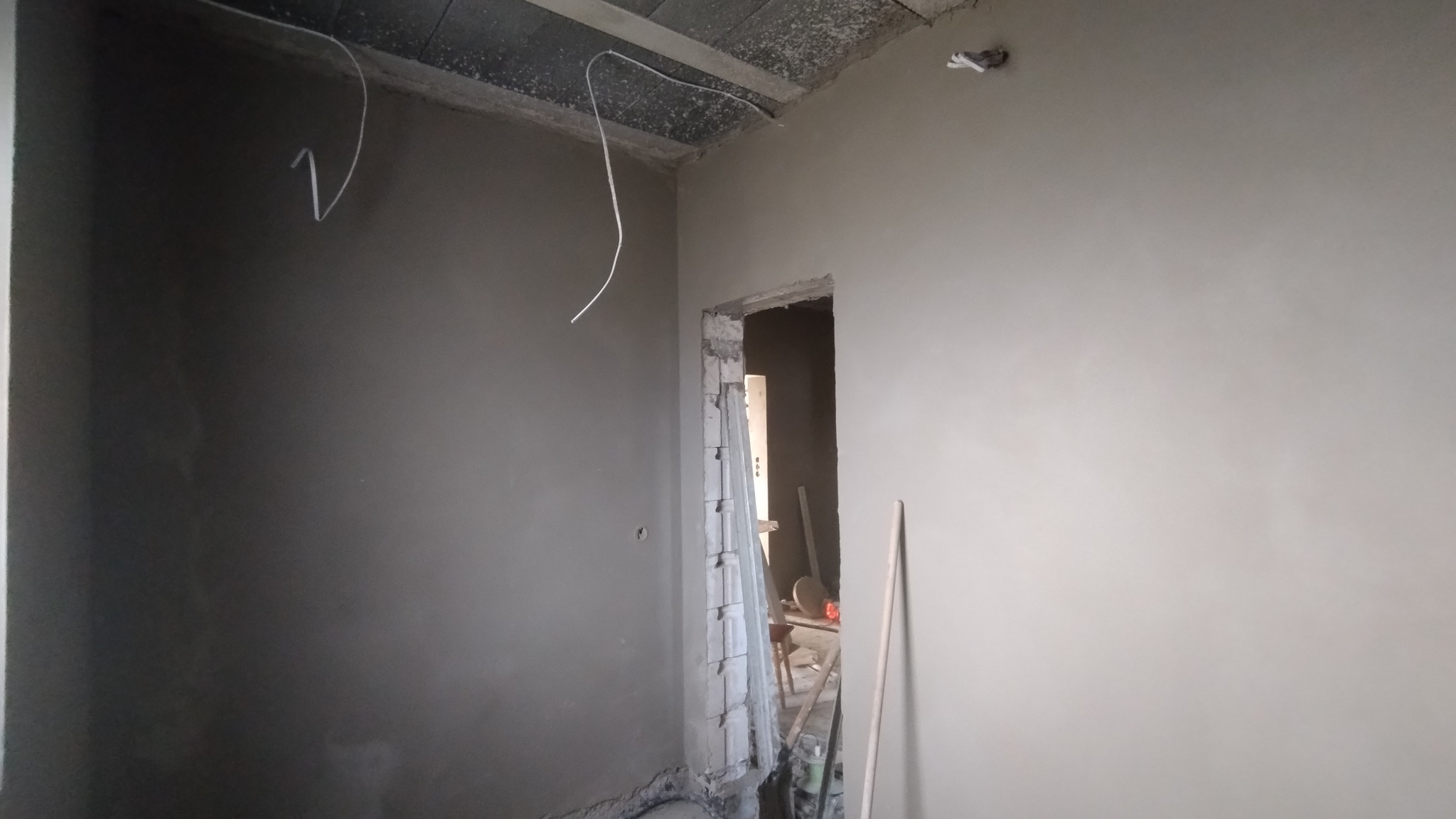 Świeżo położone tynki na ścianach wewnątrz pomieszczenia, widoczne kable wystające z sufitu i surowe obramowanie drzwi. Szare odcienie tynku.