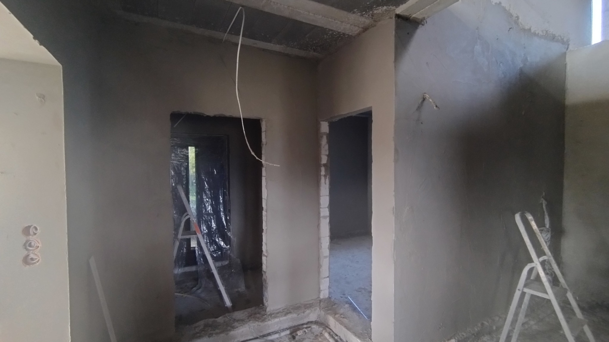 Wnętrze w trakcie tynkowania: świeżo nałożony tynk na ścianach, widoczne otwory drzwiowe, drabina oparta o ścianę. Surowy stan wykończenia.