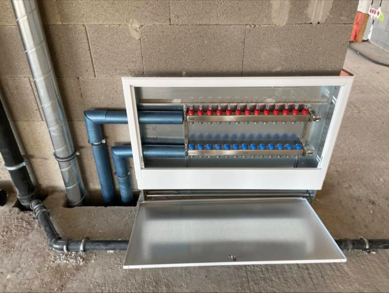 Rozdzielacz ogrzewania podłogowego w metalowej szafce, z widocznymi rurami doprowadzającymi i zaworami w kolorach czerwonym i niebieskim, na tle surowej ściany.