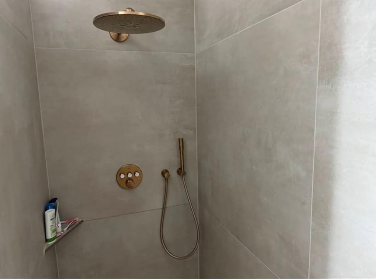 Wykończona kabina prysznicowa z deszczownicą i baterią w odcieniach złota na tle szarych, matowych płytek. Na półce kosmetyki.