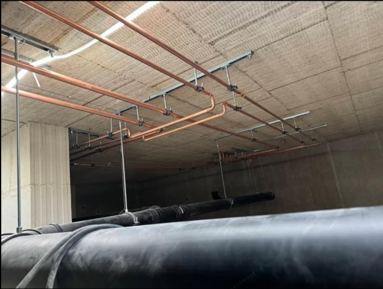 Miedziane rury instalacji CO zamocowane do sufitu na metalowych wspornikach, z widocznymi czarnymi rurami kanalizacyjnymi w dolnej części kadru.