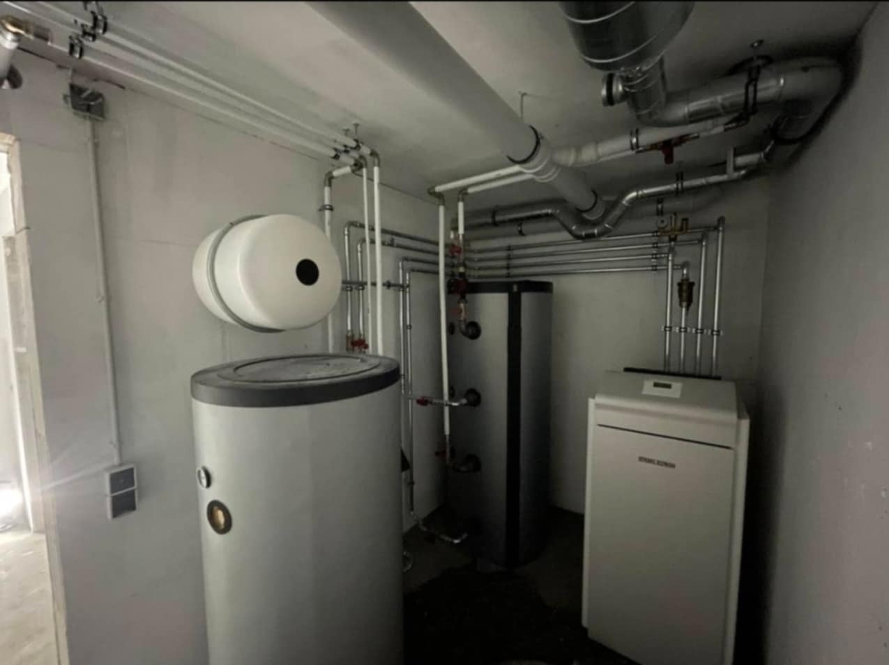 Kotłownia z trzema zbiornikami, rurami i zaworami. Widoczne różne typy instalacji grzewczych i sanitarnych w pomieszczeniu o surowym wykończeniu.