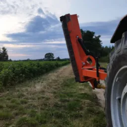 Pomarańczowy traktor z kosiarką listwową na tle zielonych krzewów i pochmurnego nieba o zmierzchu. Widoczne koło traktora i mechanizm kosiarki.