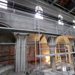 Wnętrze kościoła w trakcie remontu: rusztowania, kolumny z nałożonym tynkiem, łukowe okna w górnej części ściany, odsłonięta cegła. Prace tynkarskie w toku.