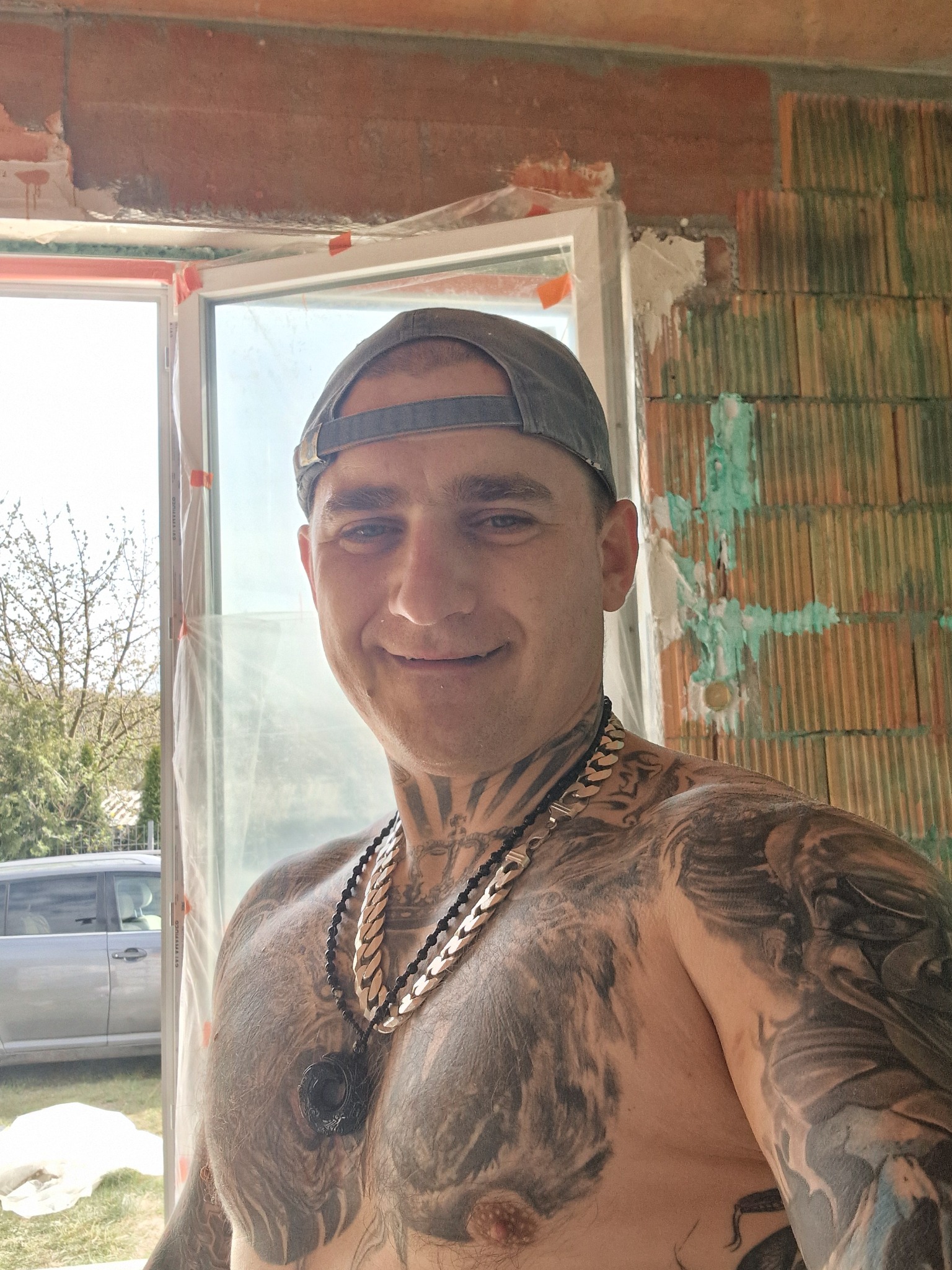 Uśmiechnięty mężczyzna w czapce, z tatuażami na tle muru z pustaków z oknem. Widoczne ślady zaprawy murarskiej i folia ochronna na ramie okiennej.
