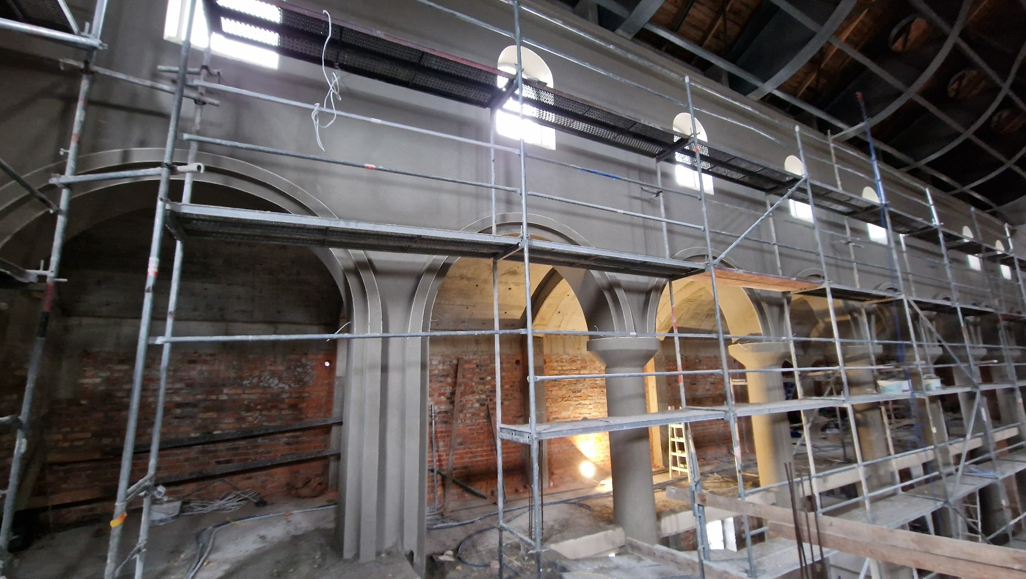 Wnętrze kościoła w trakcie remontu: rusztowania, kolumny z nałożonym tynkiem, łukowe okna w górnej części ściany, odsłonięta cegła. Prace tynkarskie w toku.