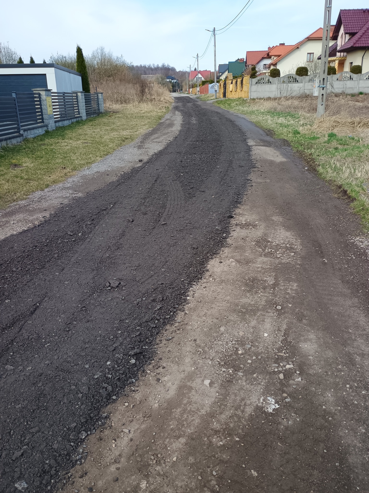 Ulepszanie drogi gruntowej poprzez wysypanie nowej warstwy. Widoczne ślady opon, okoliczne domy i trawa. Prace drogowe w toku, poprawa nawierzchni.