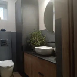 Nowoczesna łazienka z drewnianymi szafkami, umywalką nablatową i okrągłym lustrem podświetlonym od tyłu. Na blacie stoi doniczka z rośliną.