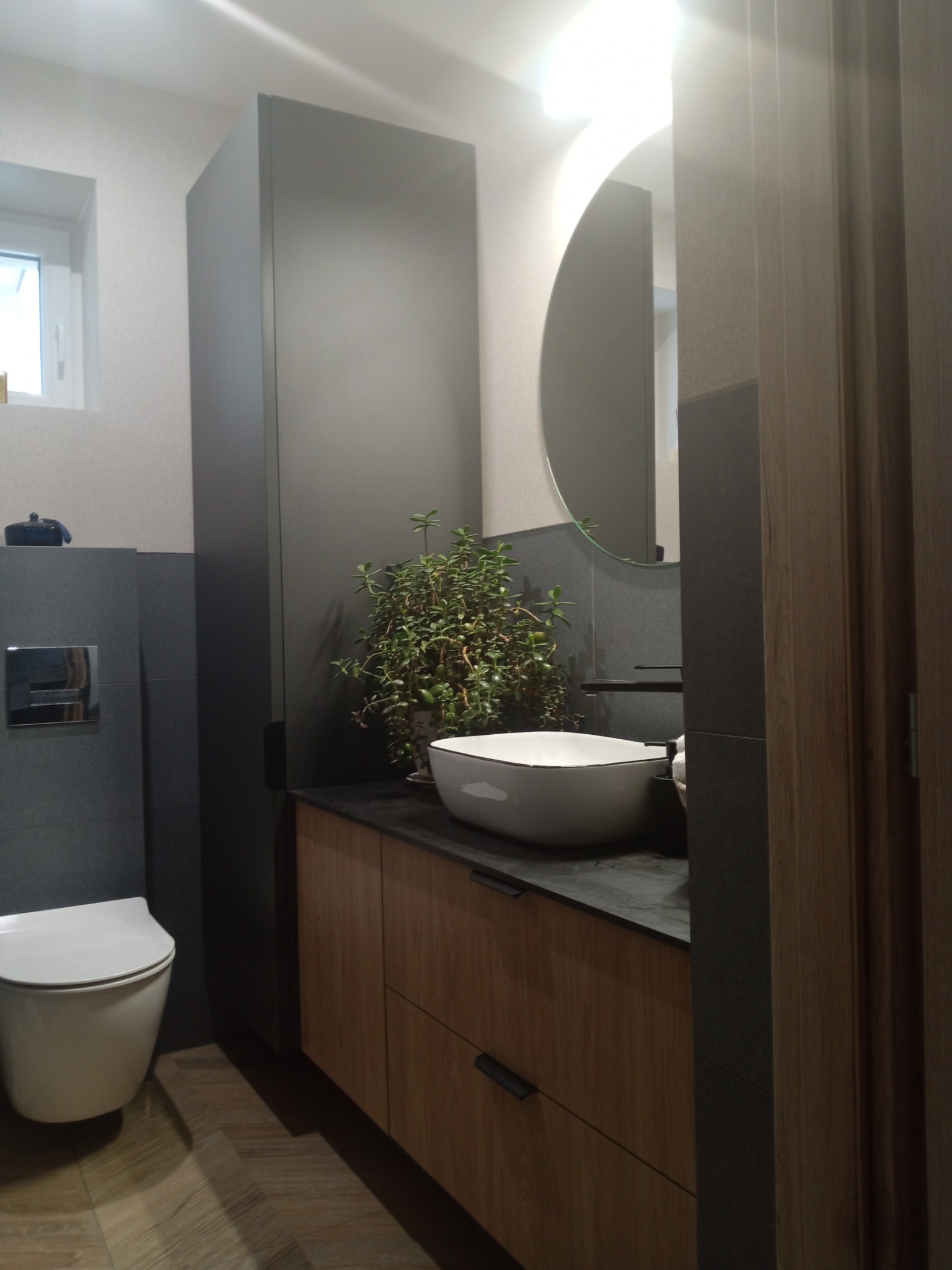 Nowoczesna łazienka z drewnianymi szafkami, umywalką nablatową i okrągłym lustrem podświetlonym od tyłu. Na blacie stoi doniczka z rośliną.