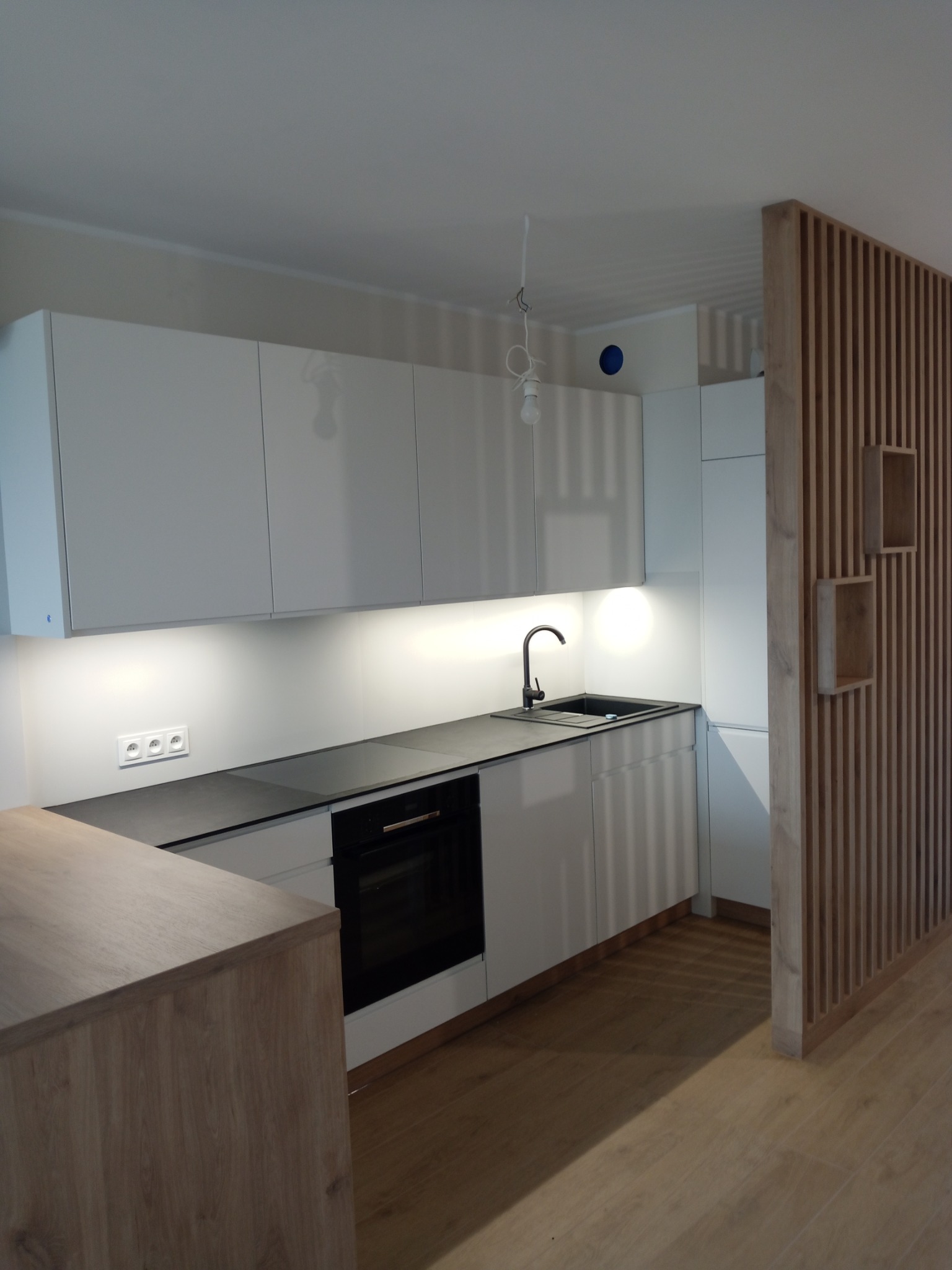 Nowoczesna, jasna kuchnia z białymi szafkami i czarnym blatem. Na podłodze panele. Drewniana ścianka działowa z półkami. Minimalistyczny design.