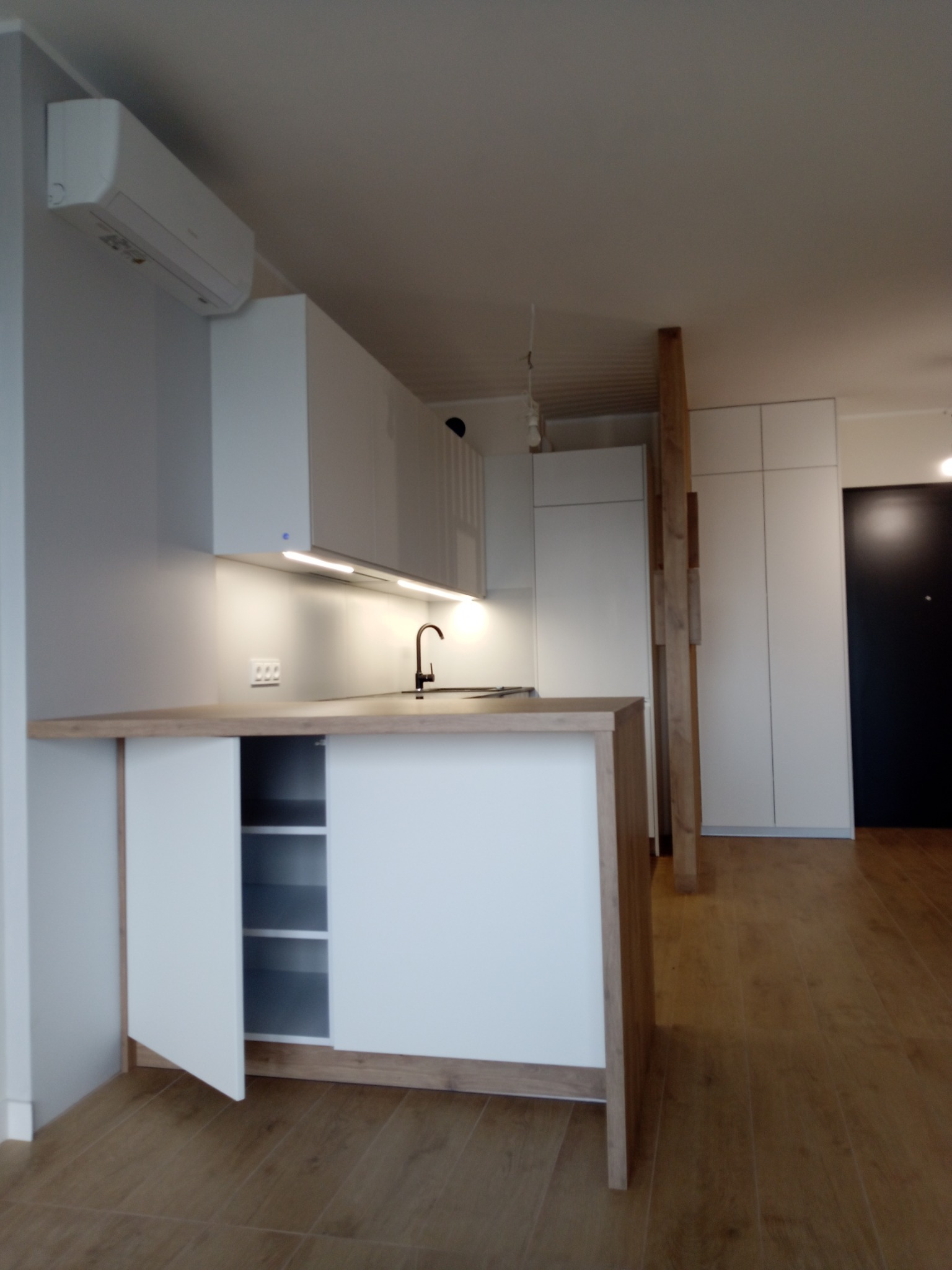 Nowoczesna, jasna kuchnia z drewnianym blatem i białymi frontami, oświetlona listwą LED. Widoczny fragment szafy wnękowej i drewniana podłoga.