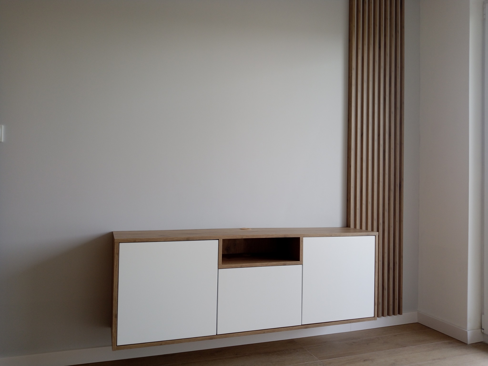 Nowoczesna szafka RTV z białymi frontami i drewnianym blatem, wisząca na szarej ścianie z dekoracyjną, pionową listwą z drewna.