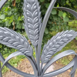 Metalowy element dekoracyjny ogrodzenia w kształcie trzech kłosów zboża, pomalowany na szaro, na tle zieleni. Detal ogrodzenia z bliska, ukazujący precyzję wykonania.