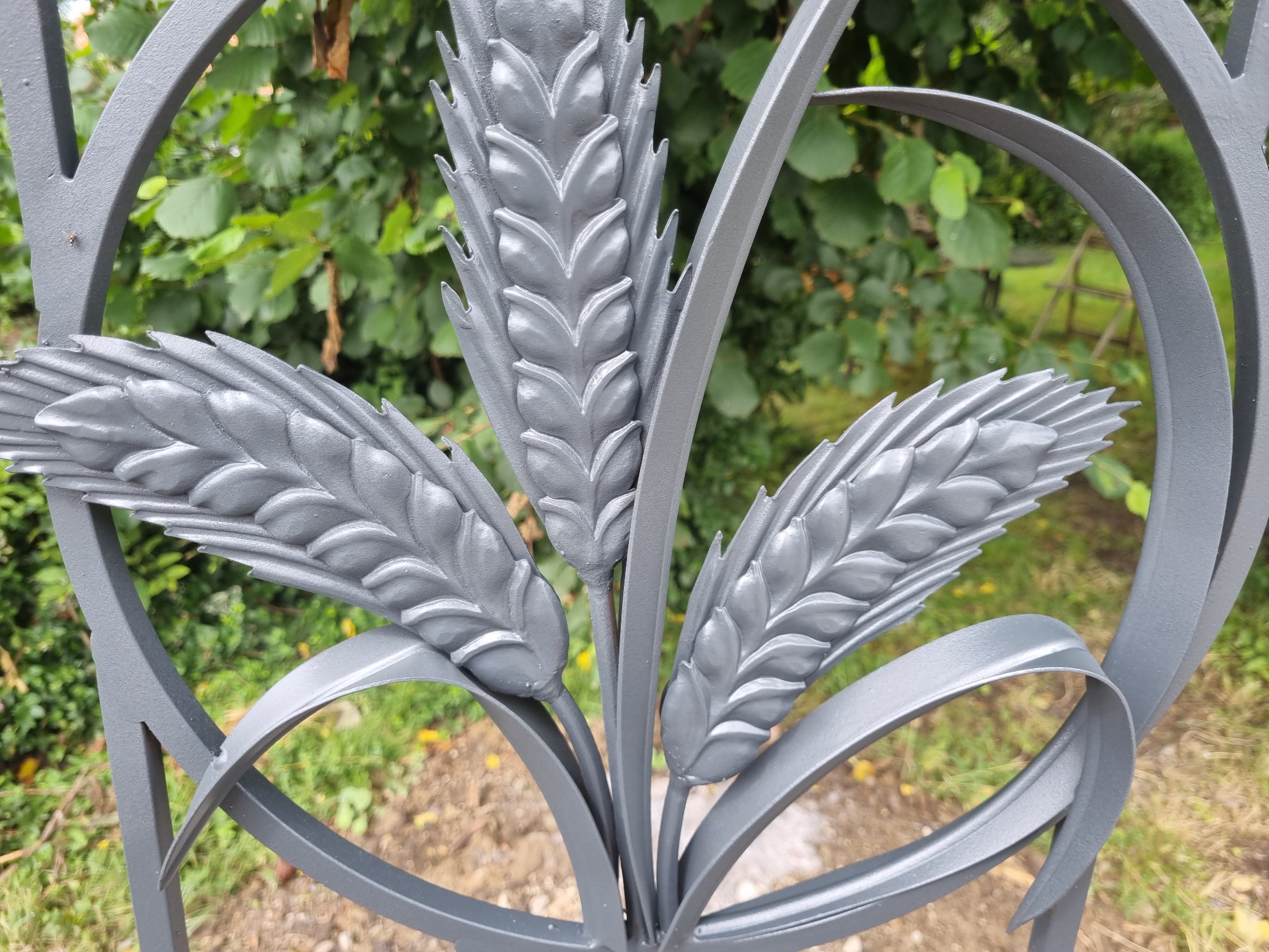 Metalowy element dekoracyjny ogrodzenia w kształcie trzech kłosów zboża, pomalowany na szaro, na tle zieleni. Detal ogrodzenia z bliska, ukazujący precyzję wykonania.