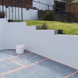 Paxon LTD - Świeżo pomalowana, biała elewacja tarasu z widocznym wiadrem farby. Tło: zielony trawnik i schody. Układ kostki z pomarańczowymi liniami.