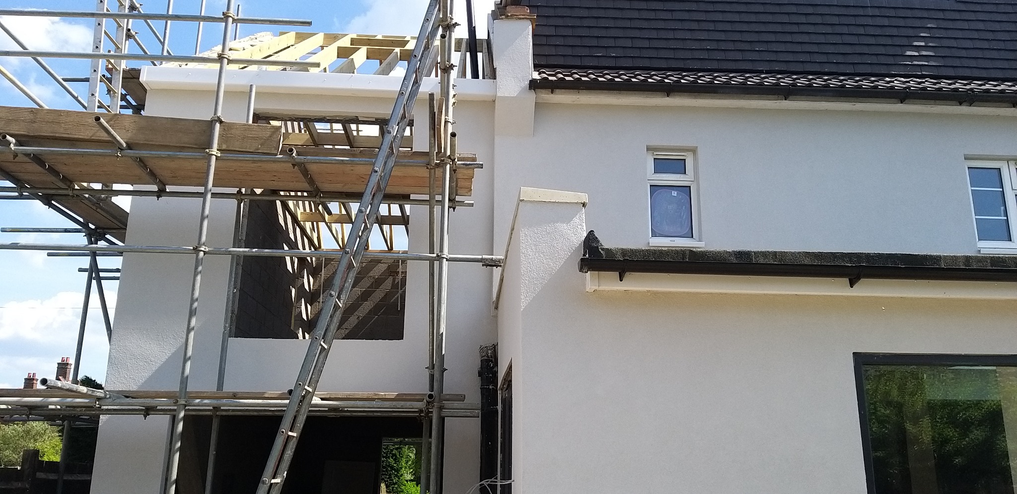Biała elewacja domu w trakcie prac wykończeniowych. Widoczne rusztowania i fragment konstrukcji dachu. Elewacja z fakturą baranka, okna PCV.