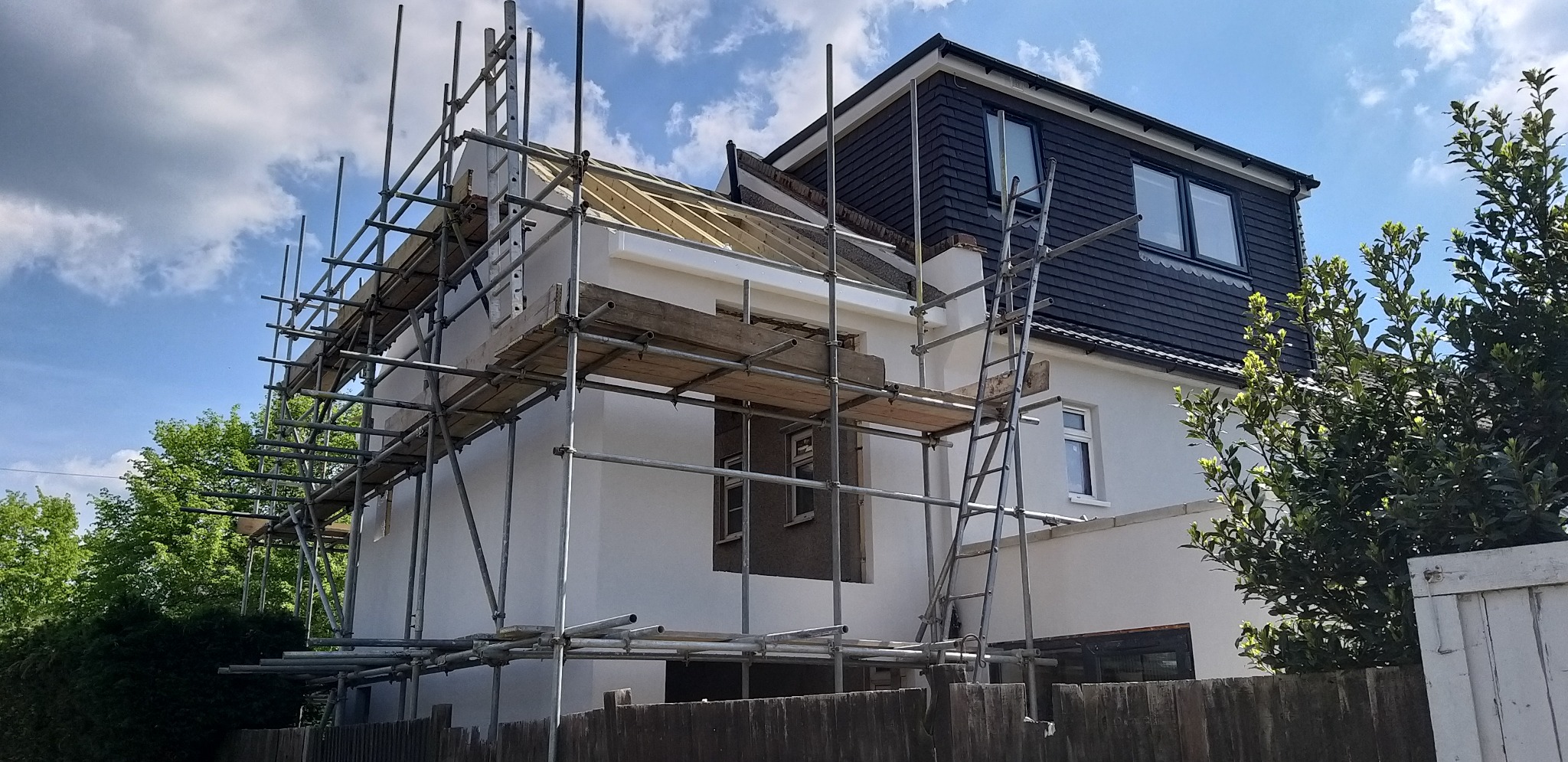 Dom w trakcie prac elewacyjnych, z widocznymi rusztowaniami i fragmentem dachu. Część fasady jest już pomalowana na biało, kontrastując z ciemniejszym fragmentem dachu.