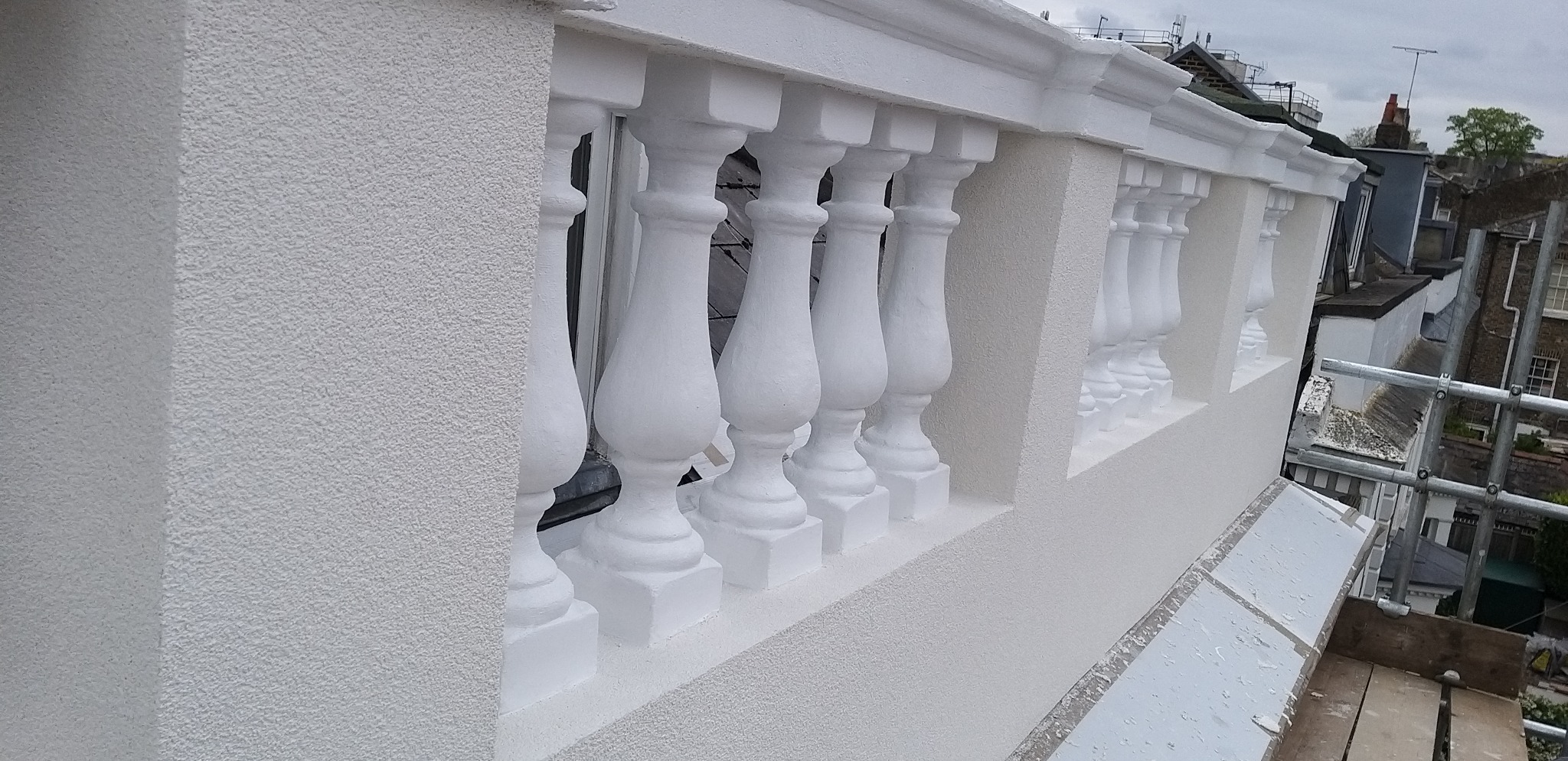 Biała elewacja z balustradą po renowacji, widok z góry. Teksturowana powierzchnia i elementy dekoracyjne. W tle zabudowa miejska i rusztowania.