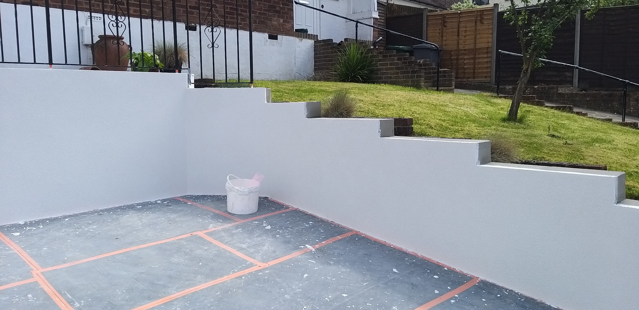 Świeżo pomalowana, biała elewacja tarasu z widocznym wiadrem farby. Tło: zielony trawnik i schody. Układ kostki z pomarańczowymi liniami.