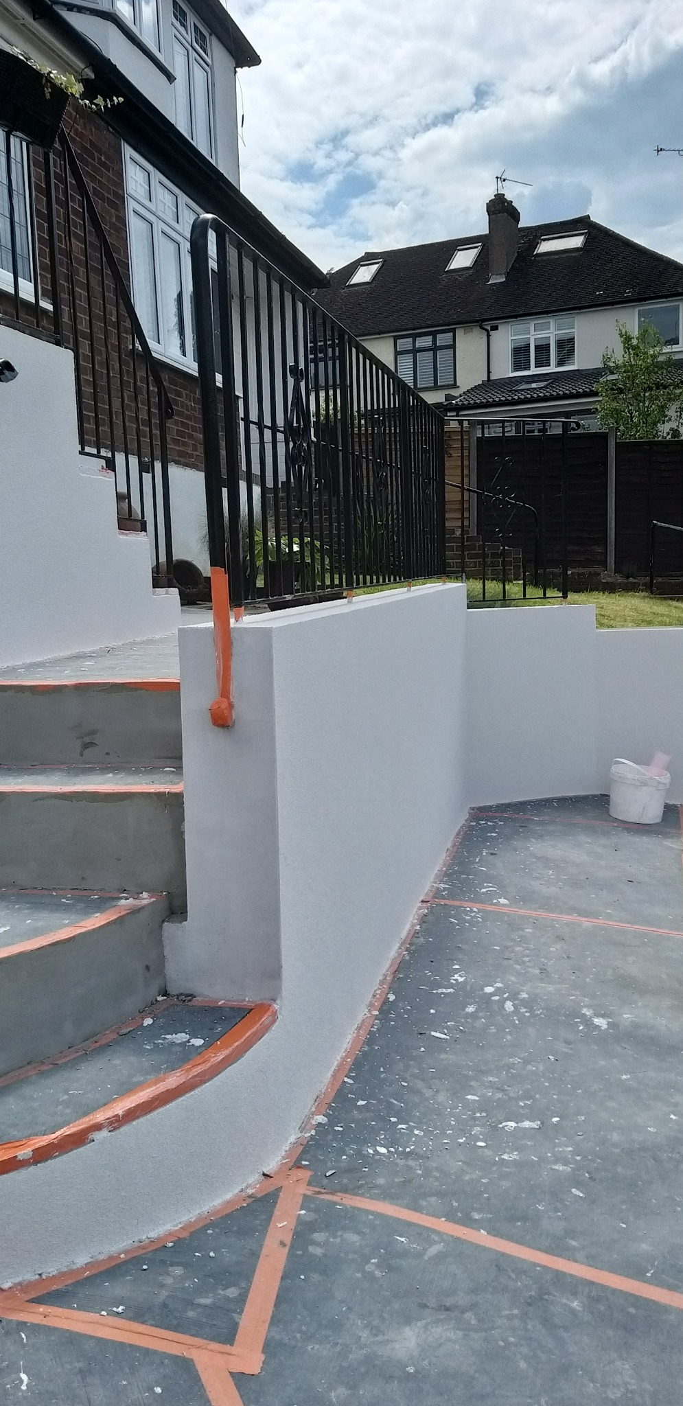 Świeżo pomalowane schody zewnętrzne i murki w jasnym odcieniu szarości, z widocznymi pomarańczowymi taśmami malarskimi i śladami farby na podłożu. Metalowa balustrada w tle.