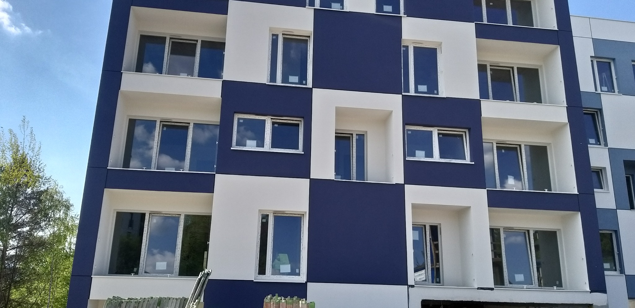 Nowoczesna elewacja budynku w szachownicę z białymi i granatowymi panelami. Okna z białymi ramami odbijają błękitne niebo. Częściowo widoczne rusztowania u dołu.