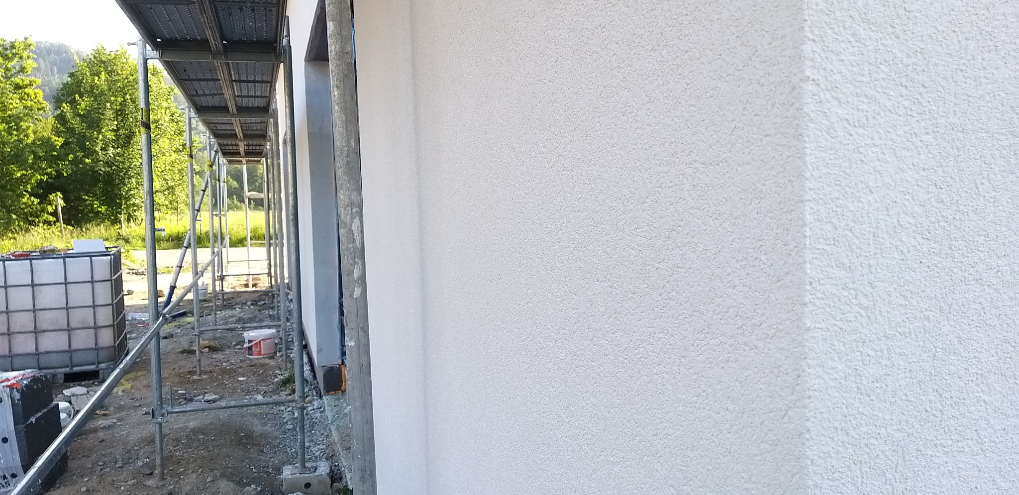 Ściana budynku po tynkowaniu, widoczne rusztowania i otoczenie. Biała elewacja z delikatną fakturą, świeżo po nałożeniu tynku. Detal wykończenia.