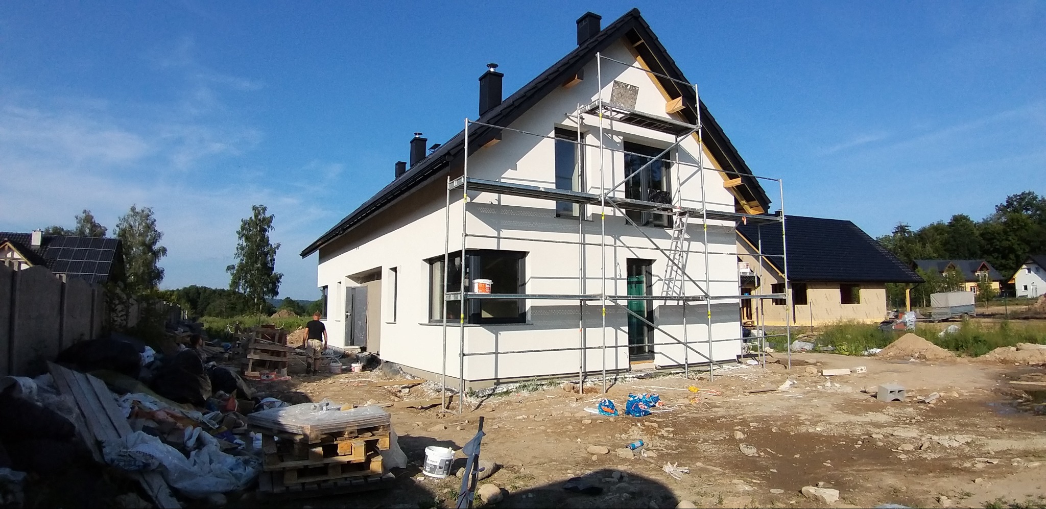 Dom jednorodzinny z nałożonym tynkiem, otoczony rusztowaniem. Widoczne detale wykończenia, czarne okna i dachówka. Ujęcie w słoneczny dzień.