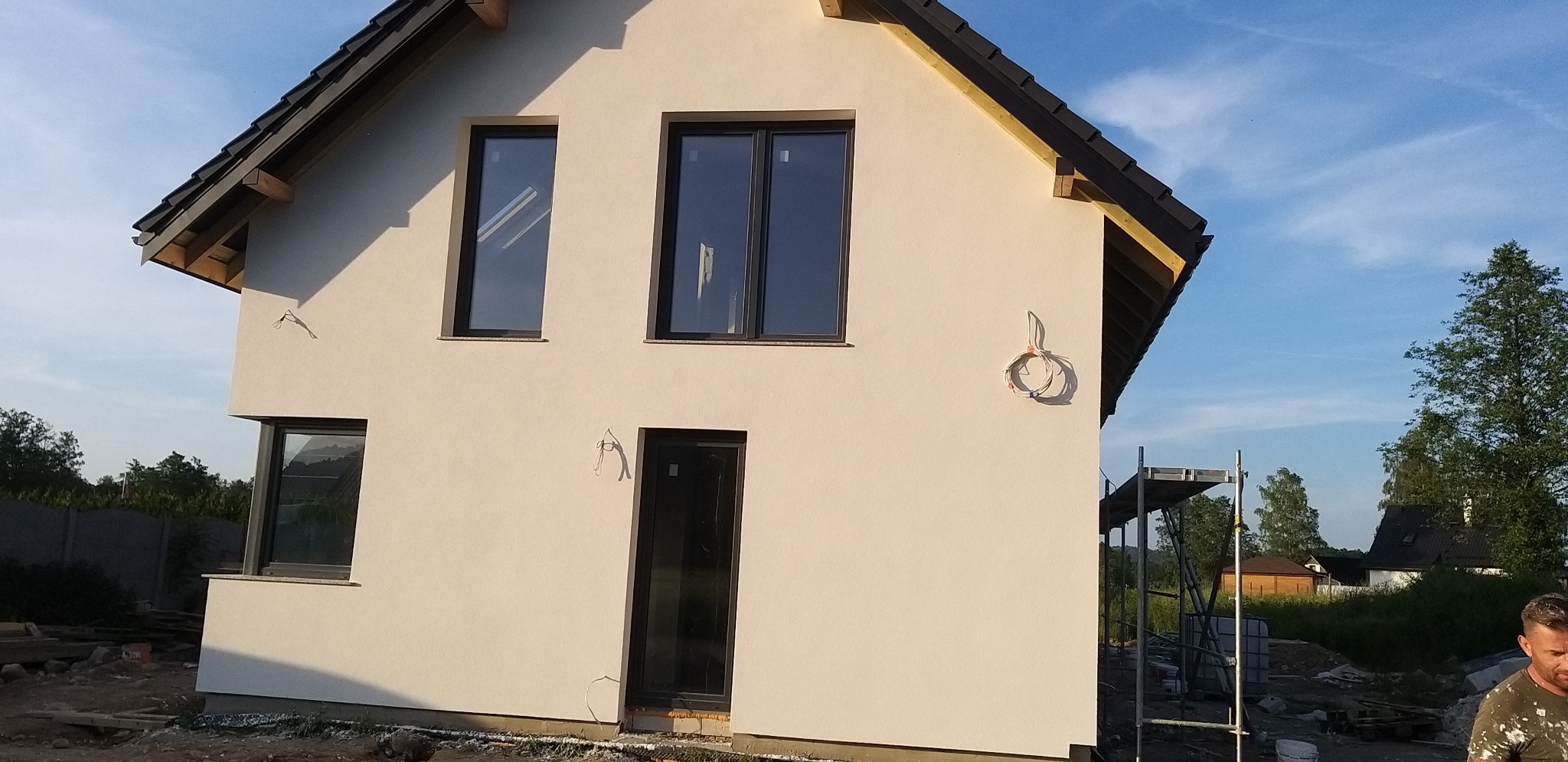 Jasna elewacja domu jednorodzinnego z czarnymi oknami i drzwiami. Na ścianie zwisający kabel. Częściowo widoczne rusztowanie i teren budowy.