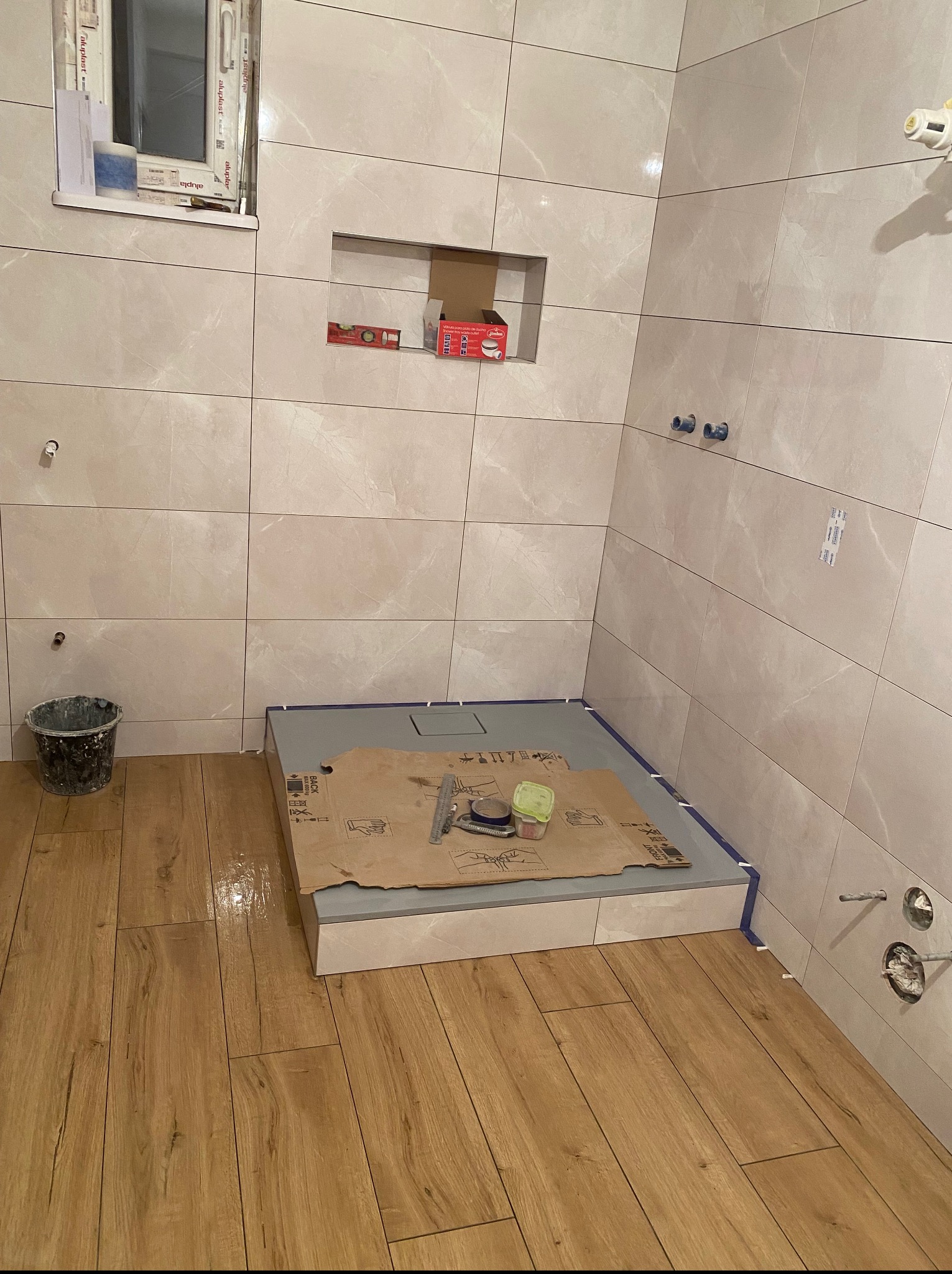 Płytki imitujące drewno na podłodze, w tle wyłożona jasnymi płytkami ściana z wnęką, przygotowana strefa prysznicowa z narzędziami.