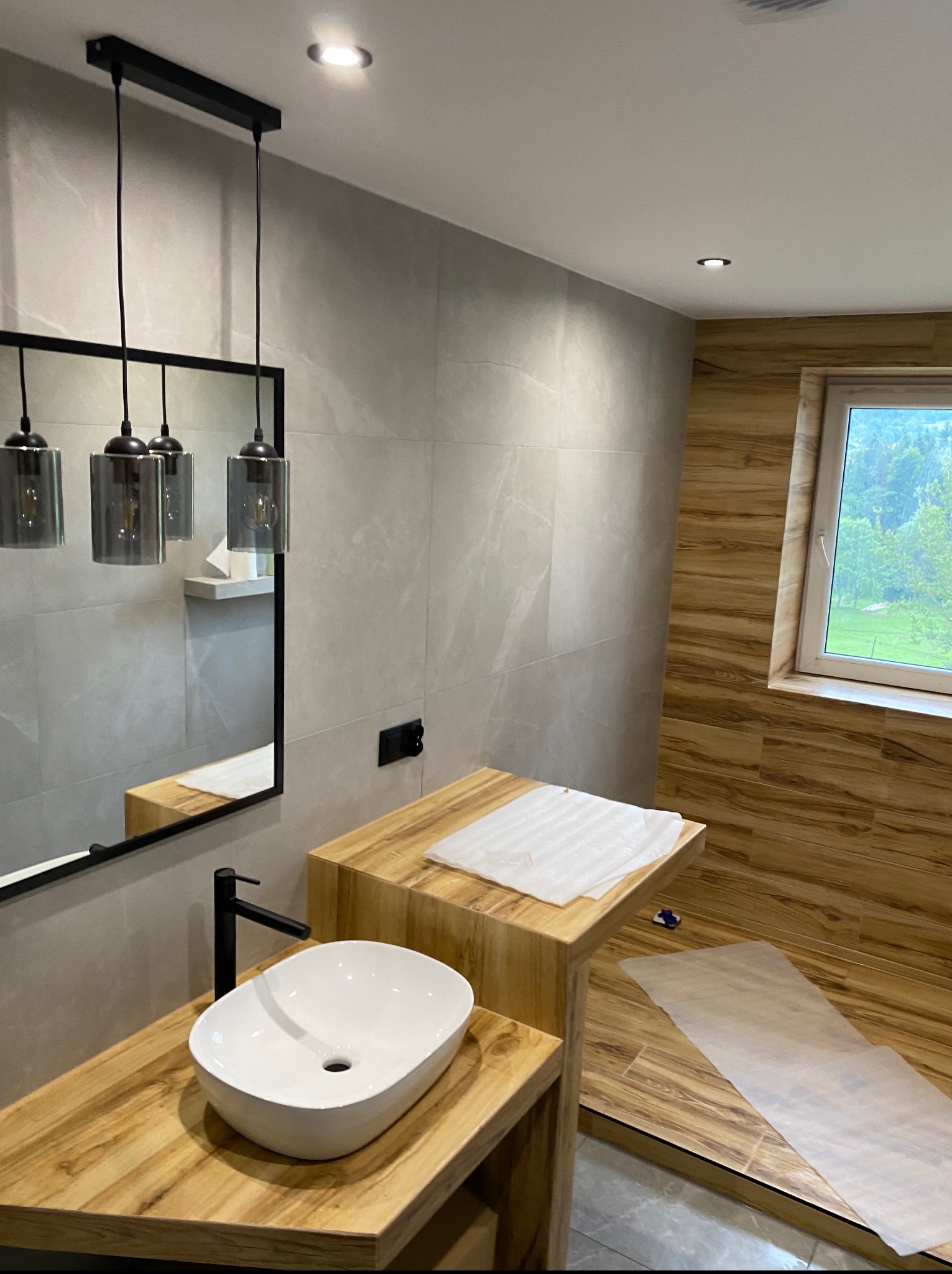 Nowoczesna łazienka z drewnianymi akcentami: blat z umywalką, lustro z lampami wiszącymi i okno z widokiem na zieleń. Płytki imitujące marmur na ścianach.