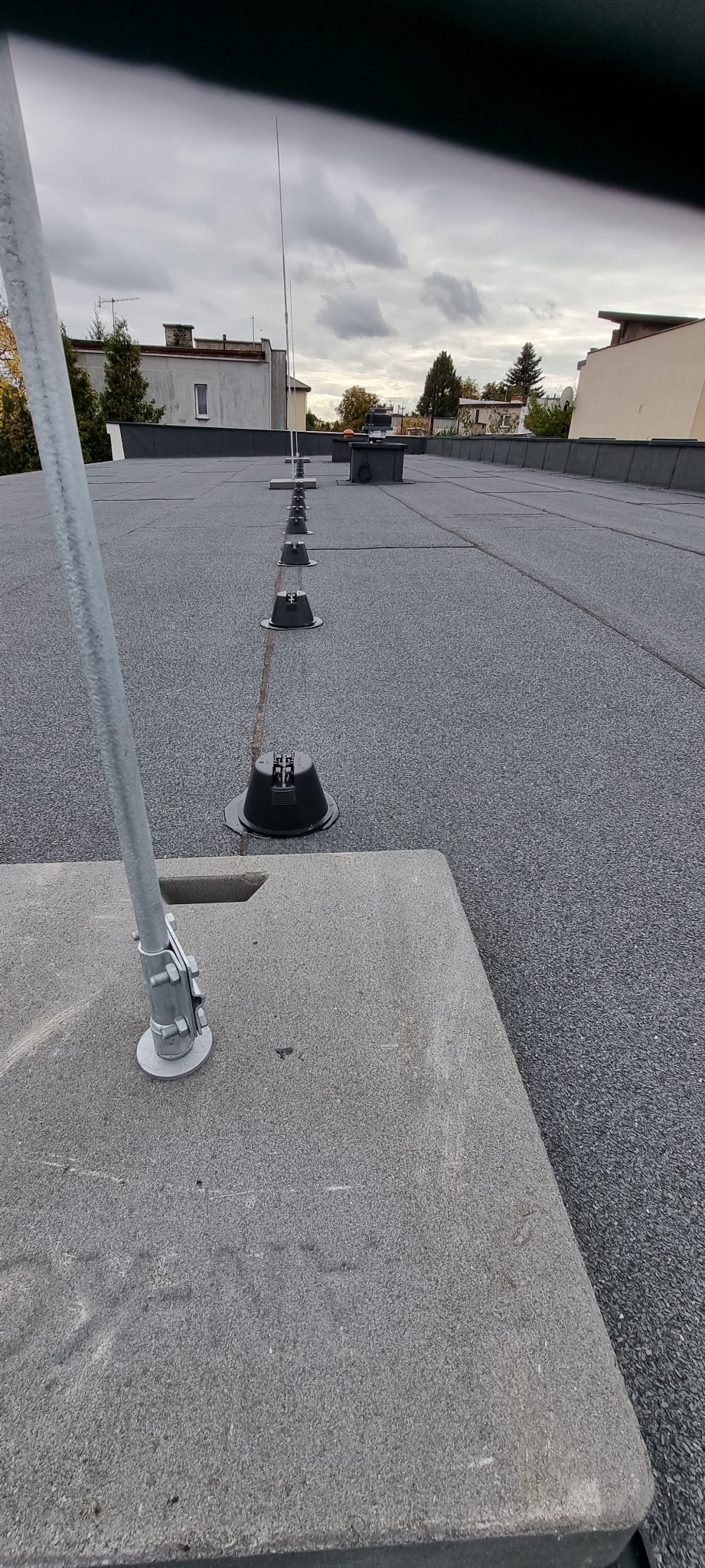 Szary dach z zamontowaną instalacją odgromową, widoczne wsporniki i maszty. Tło stanowią budynki i pochmurne niebo. Perspektywa z dołu podkreśla linię instalacji.
