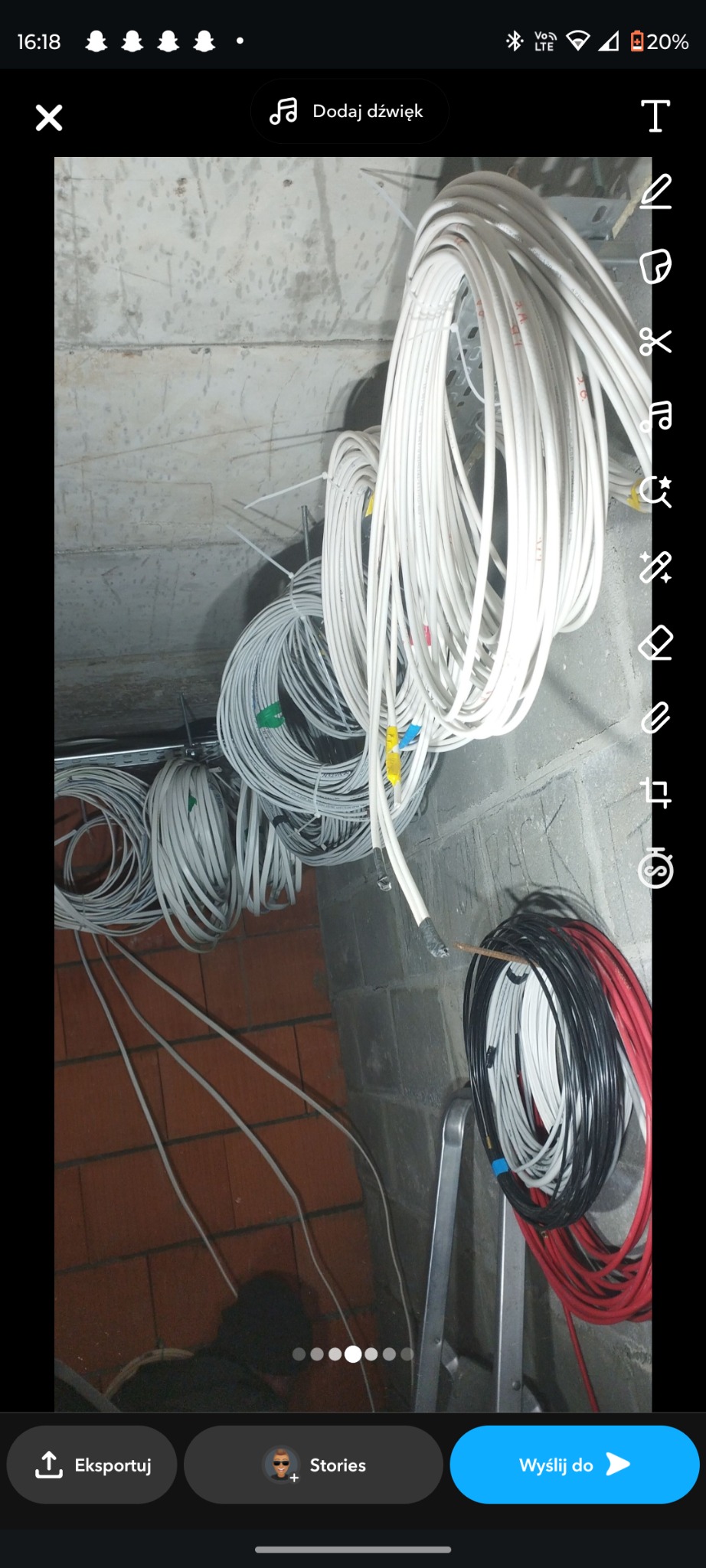 Kłębowisko przewodów elektrycznych w różnych kolorach (białe, czarne, czerwone) zwisające ze ściany w surowym stanie, z widoczną cegłą i elementami instalacji.