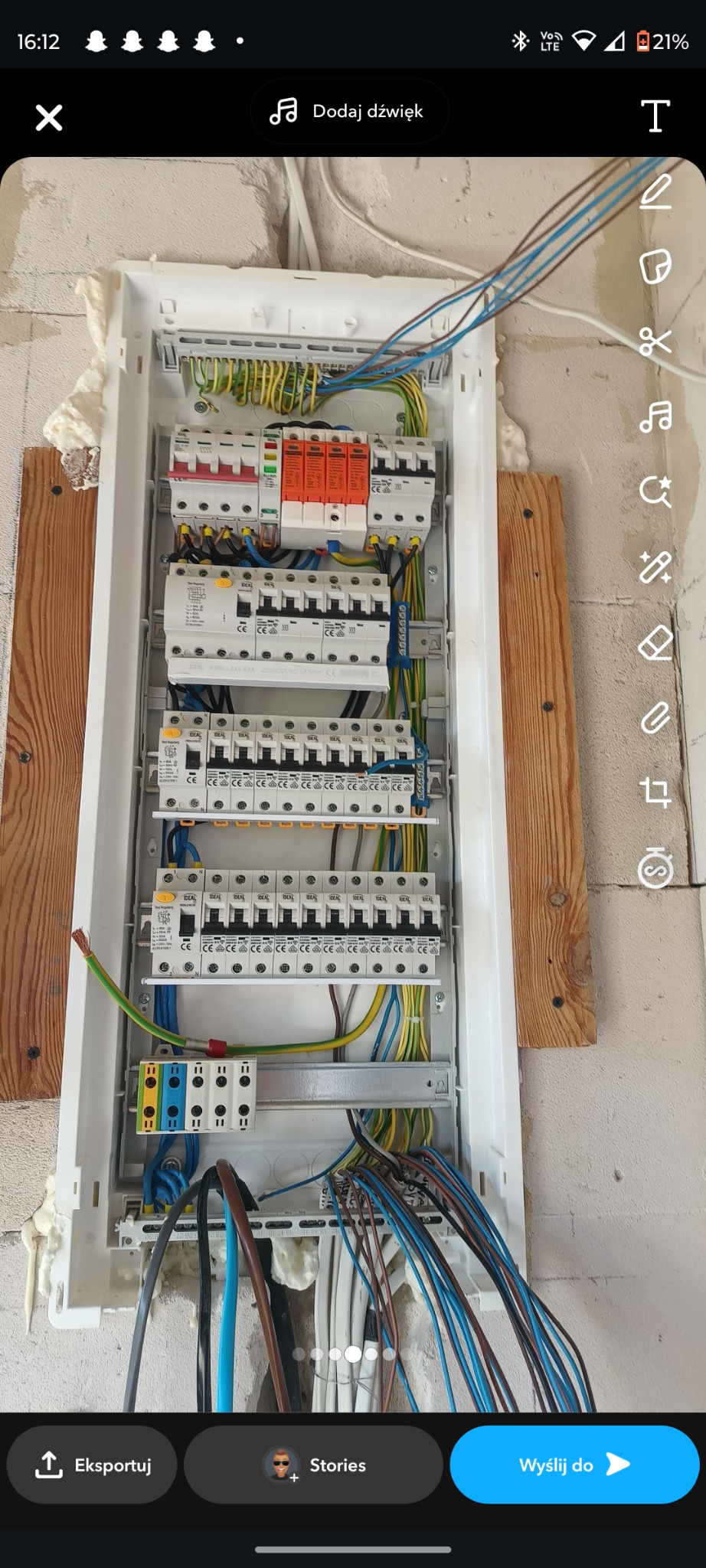 Rozdzielnia elektryczna z zamontowanymi bezpiecznikami i przewodami wewnątrz białej skrzynki, zamocowana na ścianie z drewnianymi listwami. Widoczne kable w różnych kolorach.