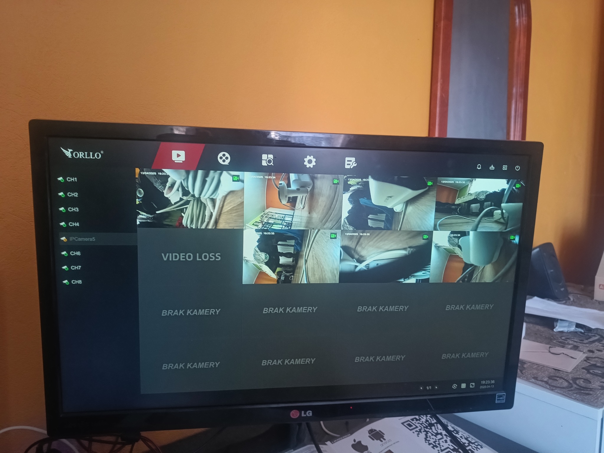 Monitor z podglądem kamer systemu monitoringu ORLLO, część kanałów niedostępna. Widoczne ustawienia, data i godzina. Pomarańczowa ściana w tle.