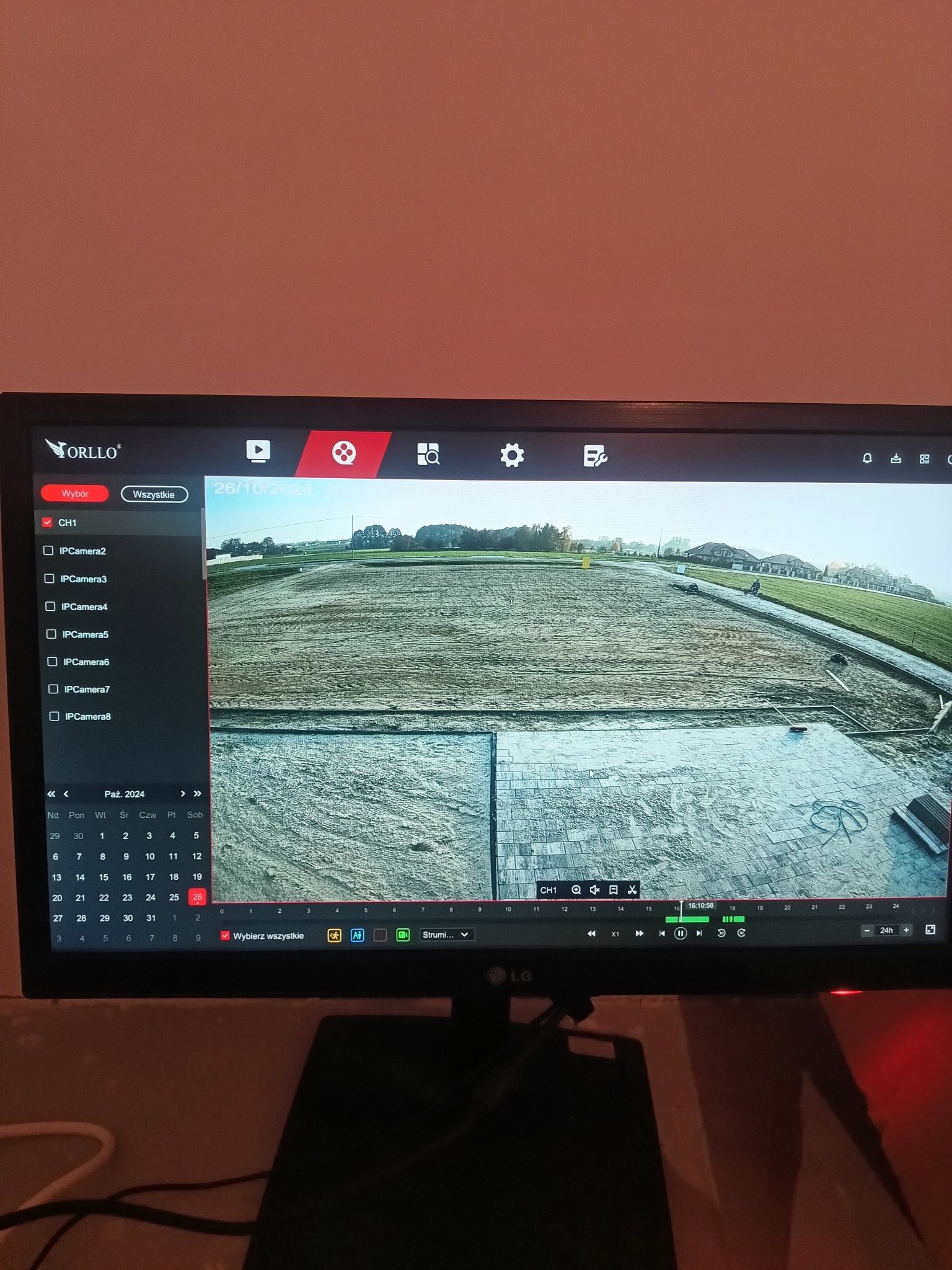 Monitor z podglądem z kamer IP Orllo, ukazujący teren budowy z kostką brukową i domami w tle, data 26.10.2024. Podgląd z systemu monitoringu.