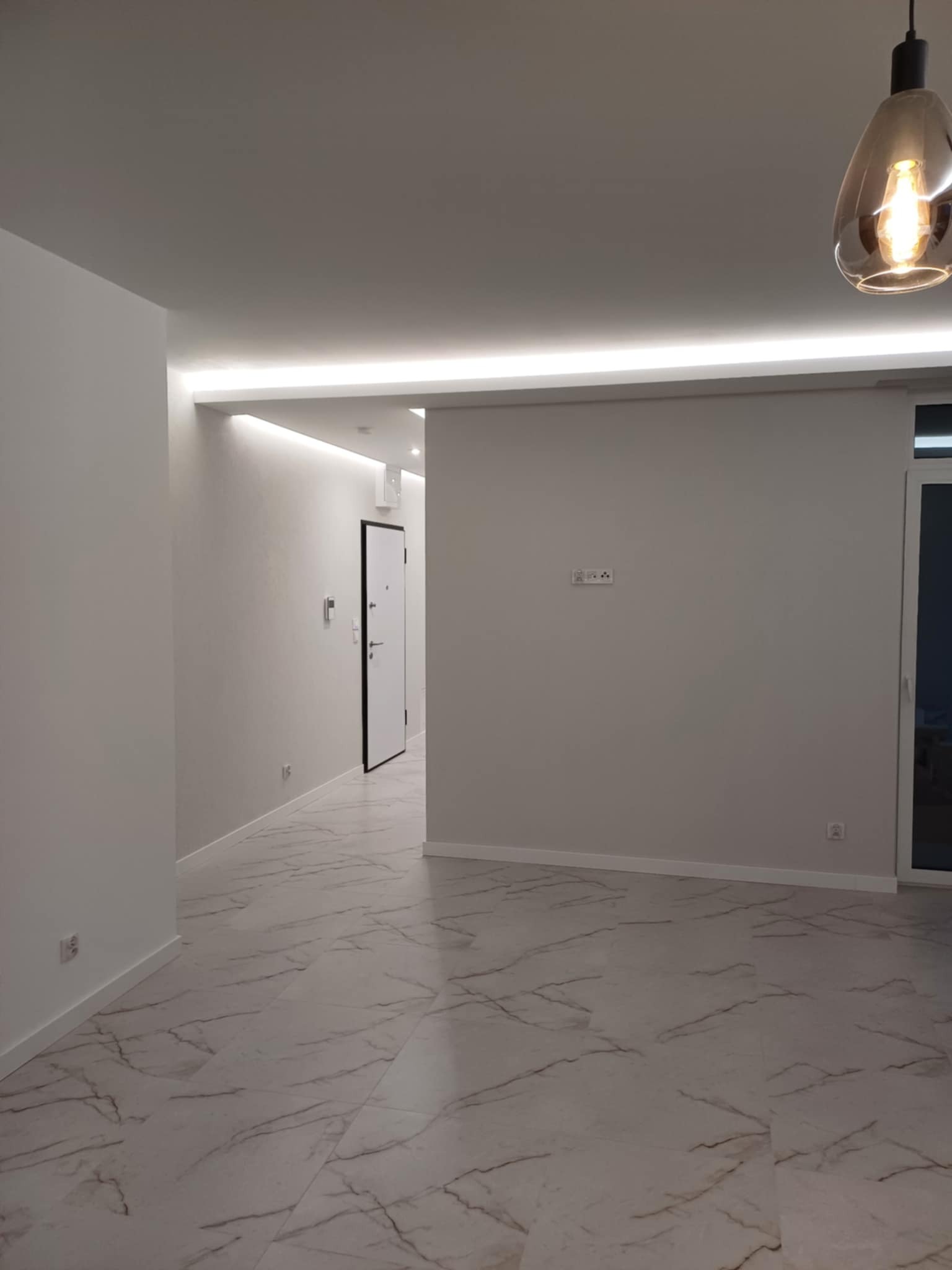 Nowoczesne wnętrze: białe ściany, marmurowa podłoga, oświetlenie LED w suficie i wisząca lampa. Widoczne gniazdka elektryczne i drzwi wejściowe.