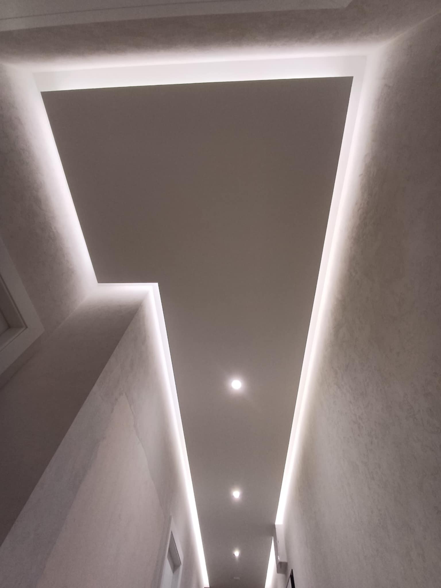 Nowoczesny sufit z oświetleniem LED w kształcie litery V, punktowe lampy, jasne ściany z delikatną fakturą. Minimalistyczny design wnętrza.