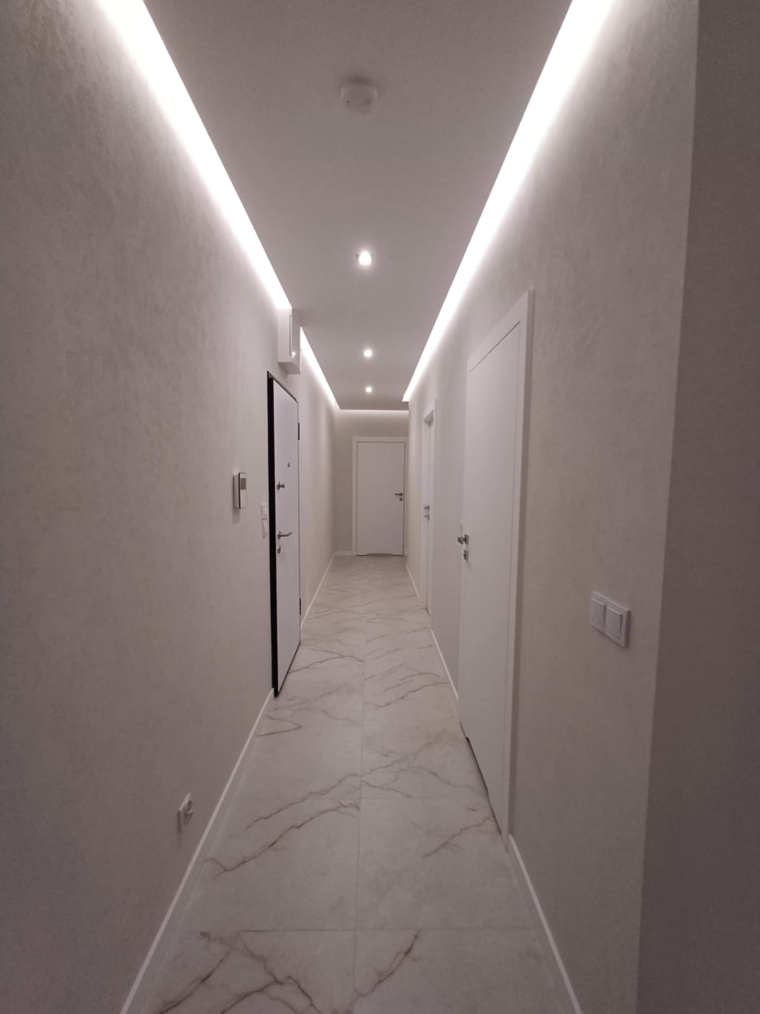 Nowoczesny korytarz z oświetleniem LED wzdłuż sufitu, marmurowa podłoga i białe drzwi. Minimalistyczny design z subtelnym oświetleniem punktowym.