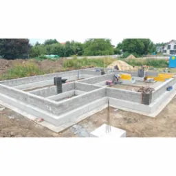 Fundamenty z bloczków betonowych, zbrojenie pionowe, szalunki. Widok na plac budowy z zielenią w tle. Prace fundamentowe w toku, widoczny pracownik i sprzęt budowlany.