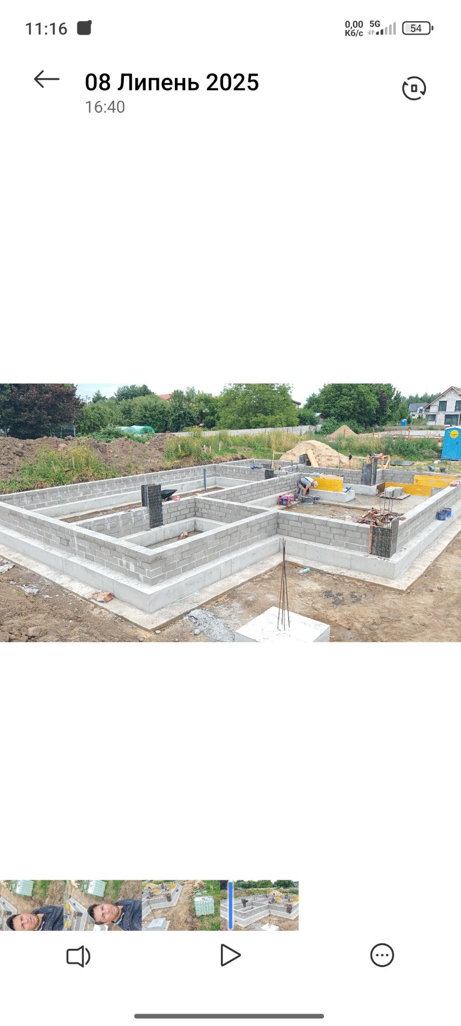 Fundamenty z bloczków betonowych, zbrojenie pionowe, szalunki. Widok na plac budowy z zielenią w tle. Prace fundamentowe w toku, widoczny pracownik i sprzęt budowlany.